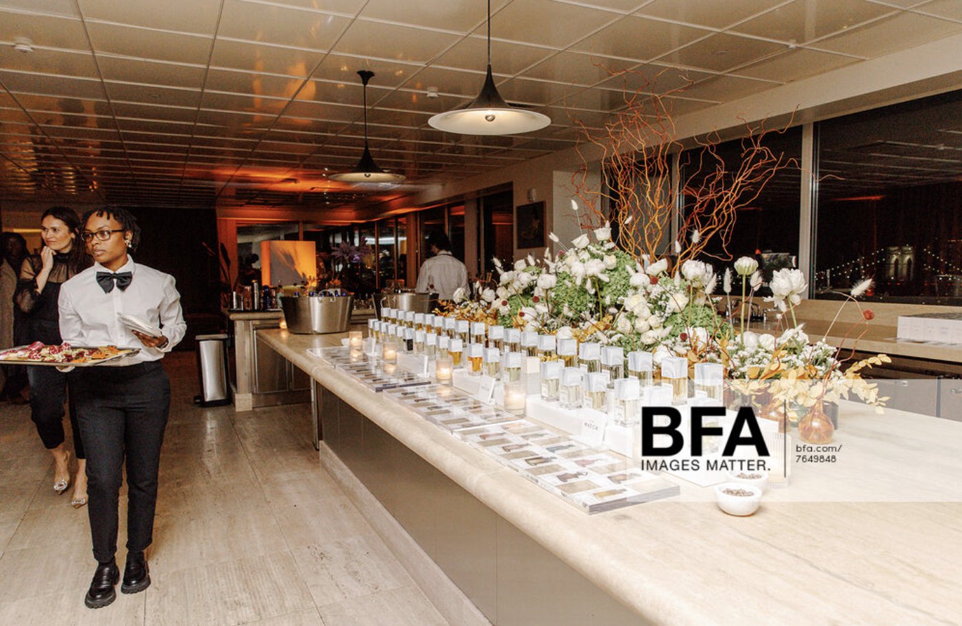 A waitress in a white shirt and black pants carrying a tray with food at a formal event, with a large floral arrangement and numerous small candles or containers on a table beside her.