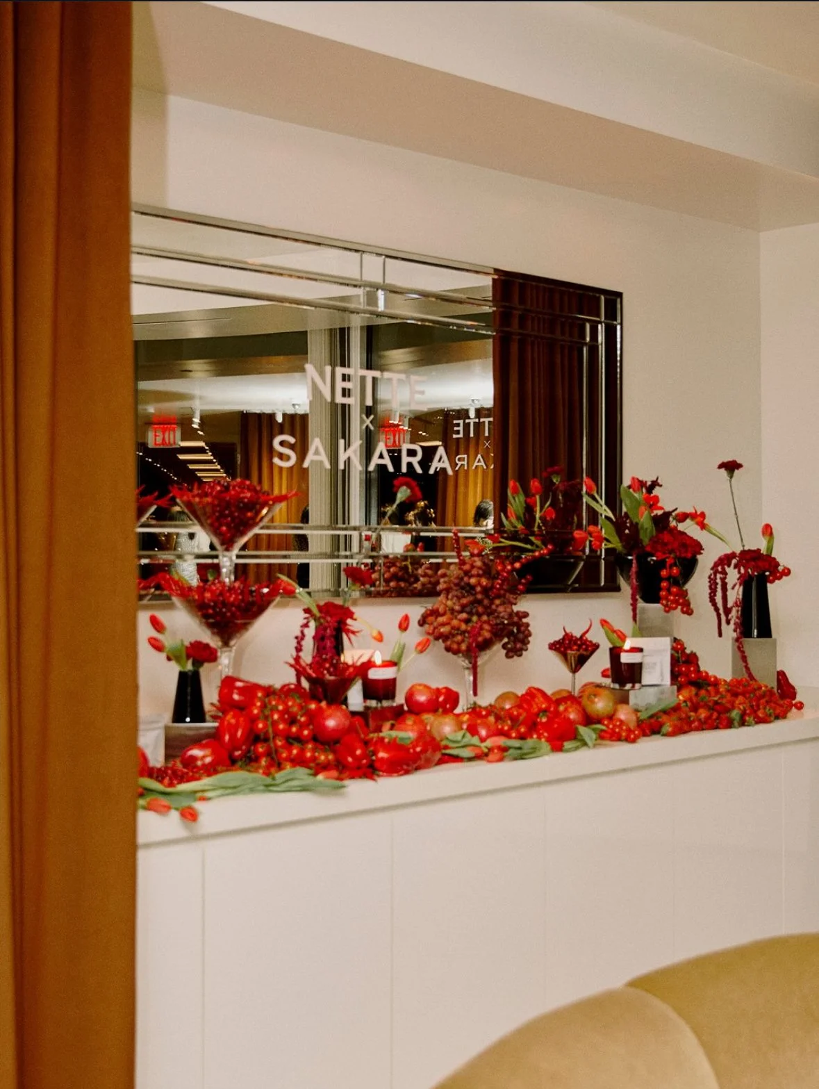 Decorative display of red fruits, berries, and flowers on a white countertop with vases and glasses, reflection of a room with wooden curtains and signage in the mirror.