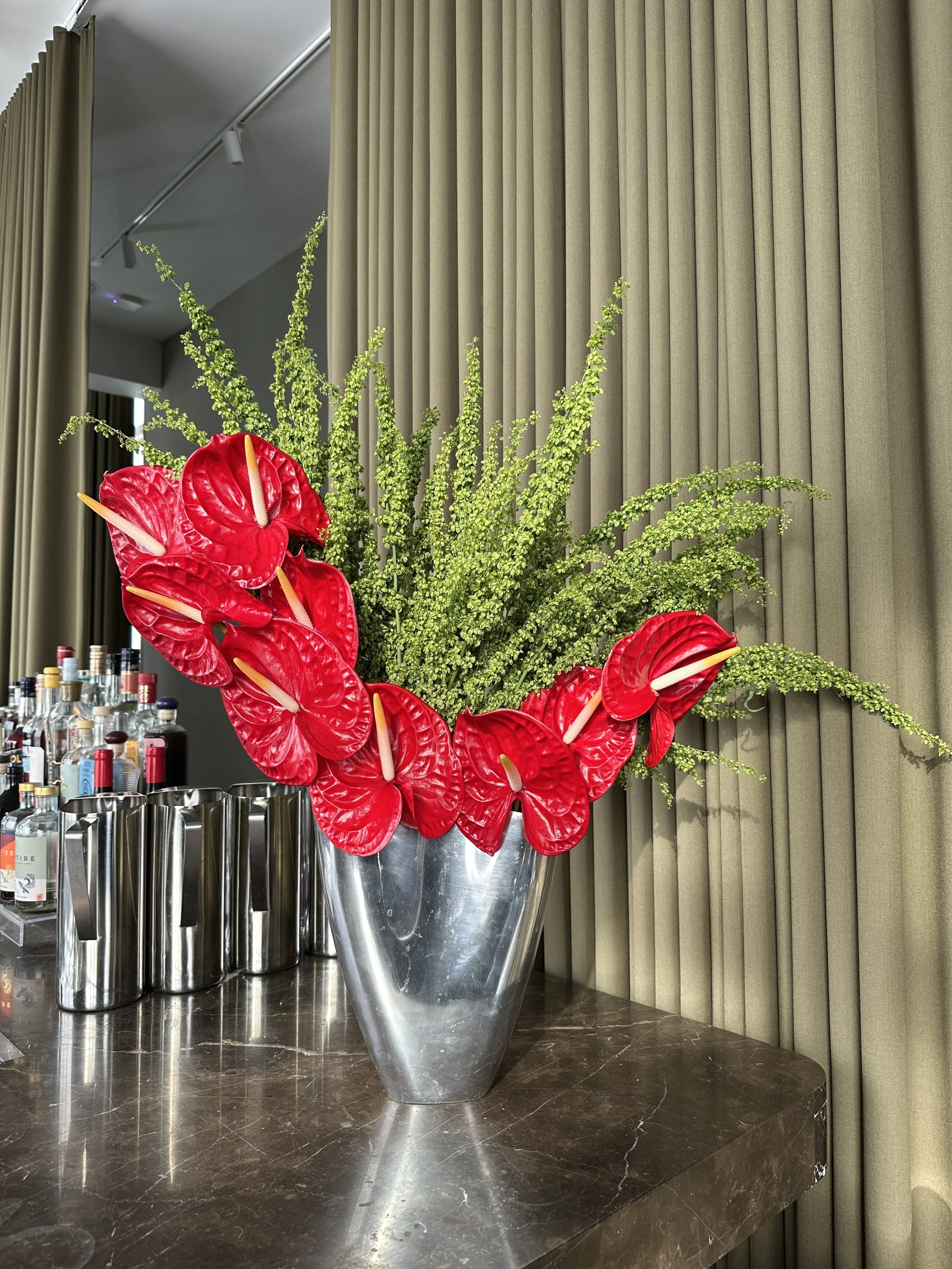 Decorative arrangement of red anthurium flowers and green foliage in a metallic vase, placed on a marble countertop near a bar with liquor bottles.