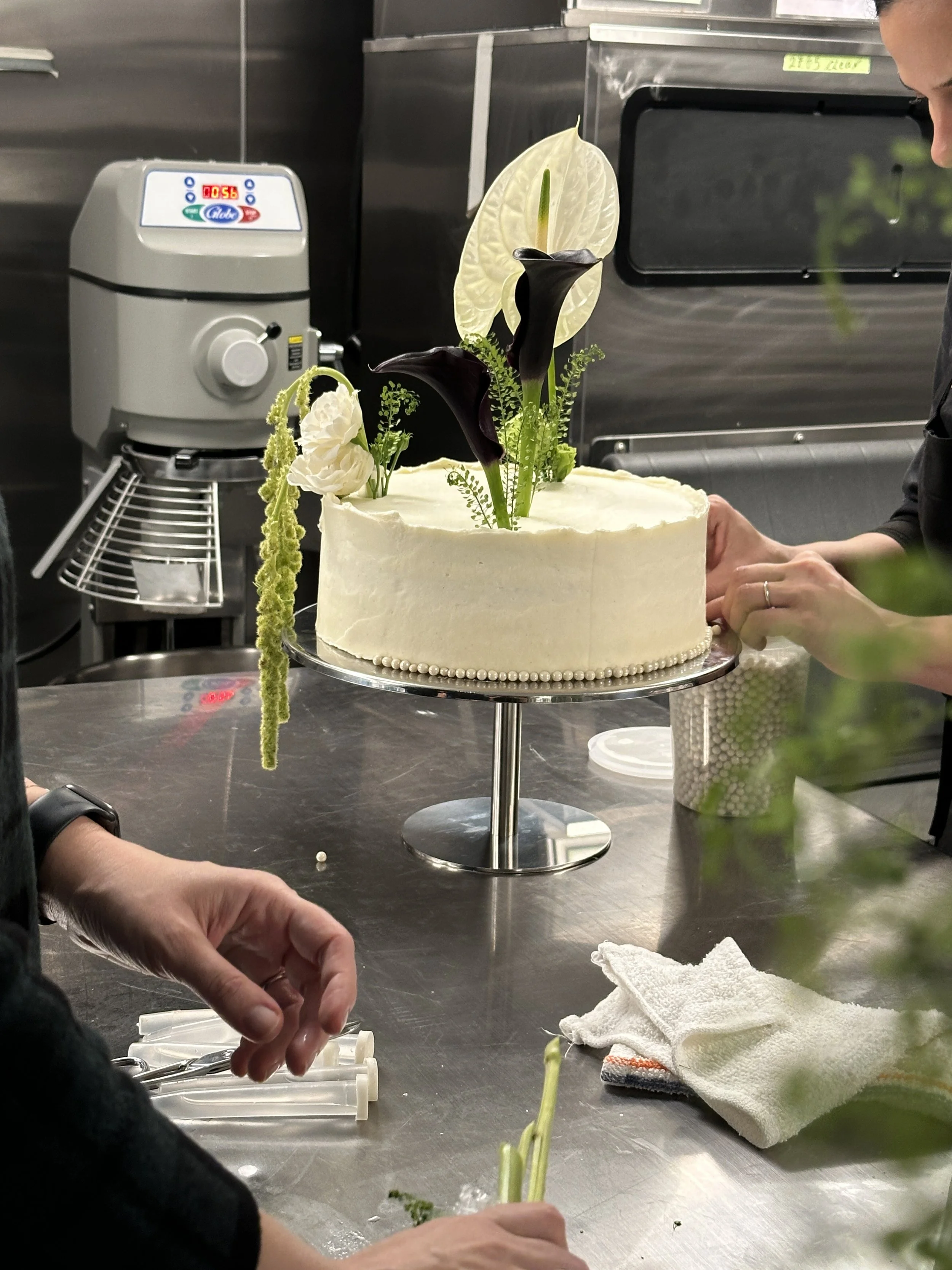 A decorated white cake with black and white flowers on top, displayed on a cake stand in a kitchen.
