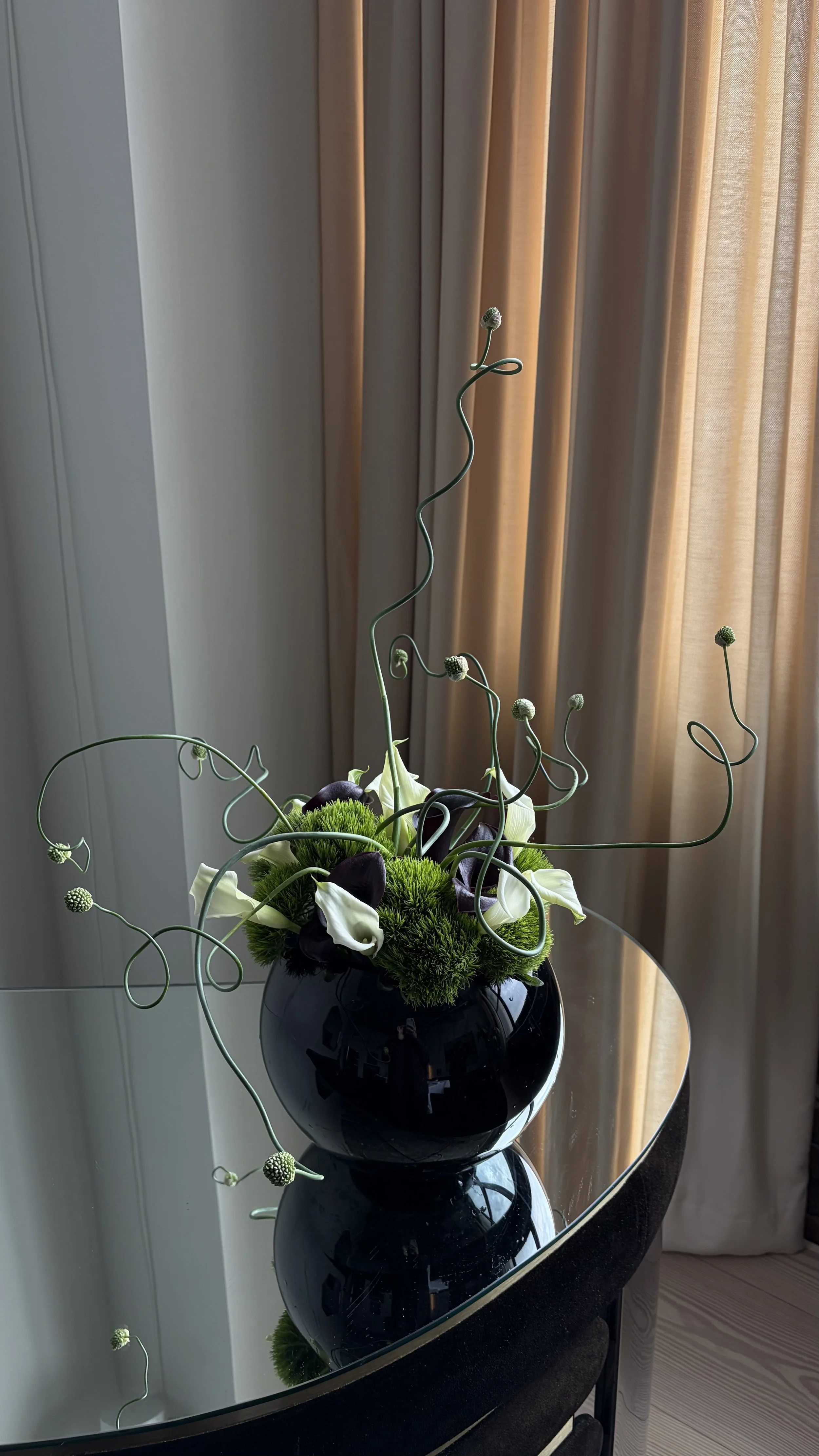 A black vase with white calla lilies, dark purple flowers, green moss, and curly decorative twigs on a mirrored table near beige curtains.