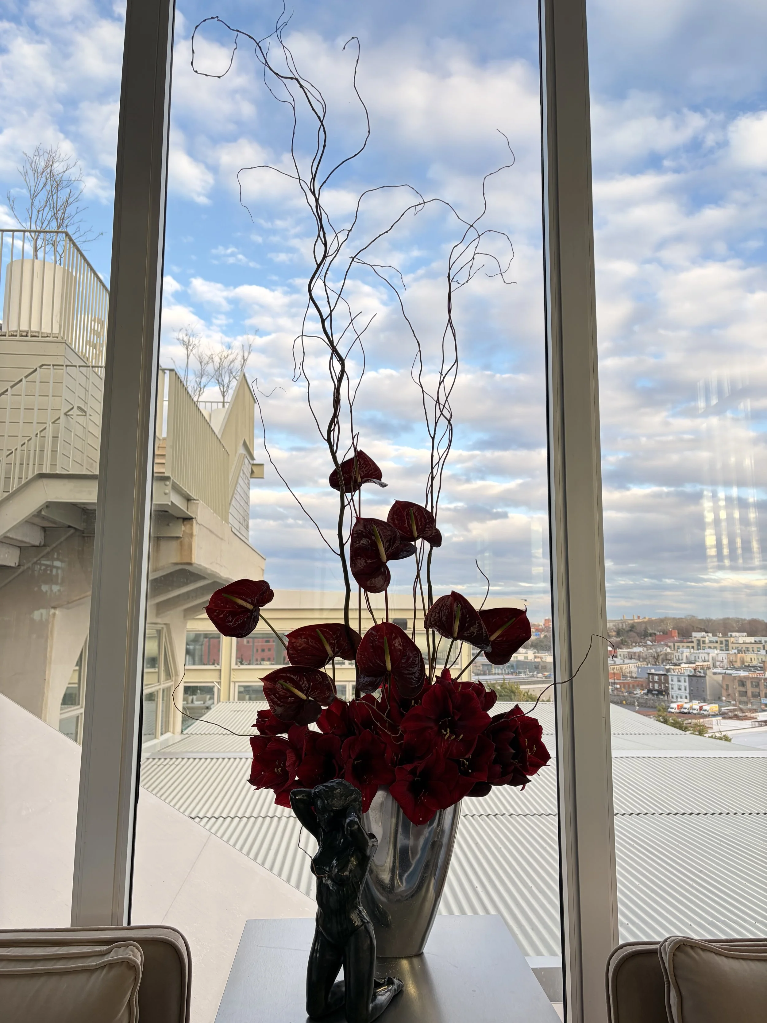 A tall, decorative plant with large, dark red flowers and curly, twig-like branches in a metallic vase, placed on a table near a large window with a cityscape view and blue sky with clouds in the background.