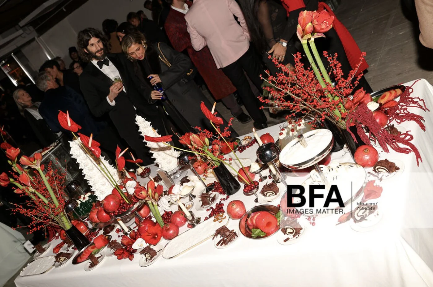 A formal event with a table decorated in red and white flowers, pomegranates, and chocolate desserts, with guests dressed in evening attire observing the display.