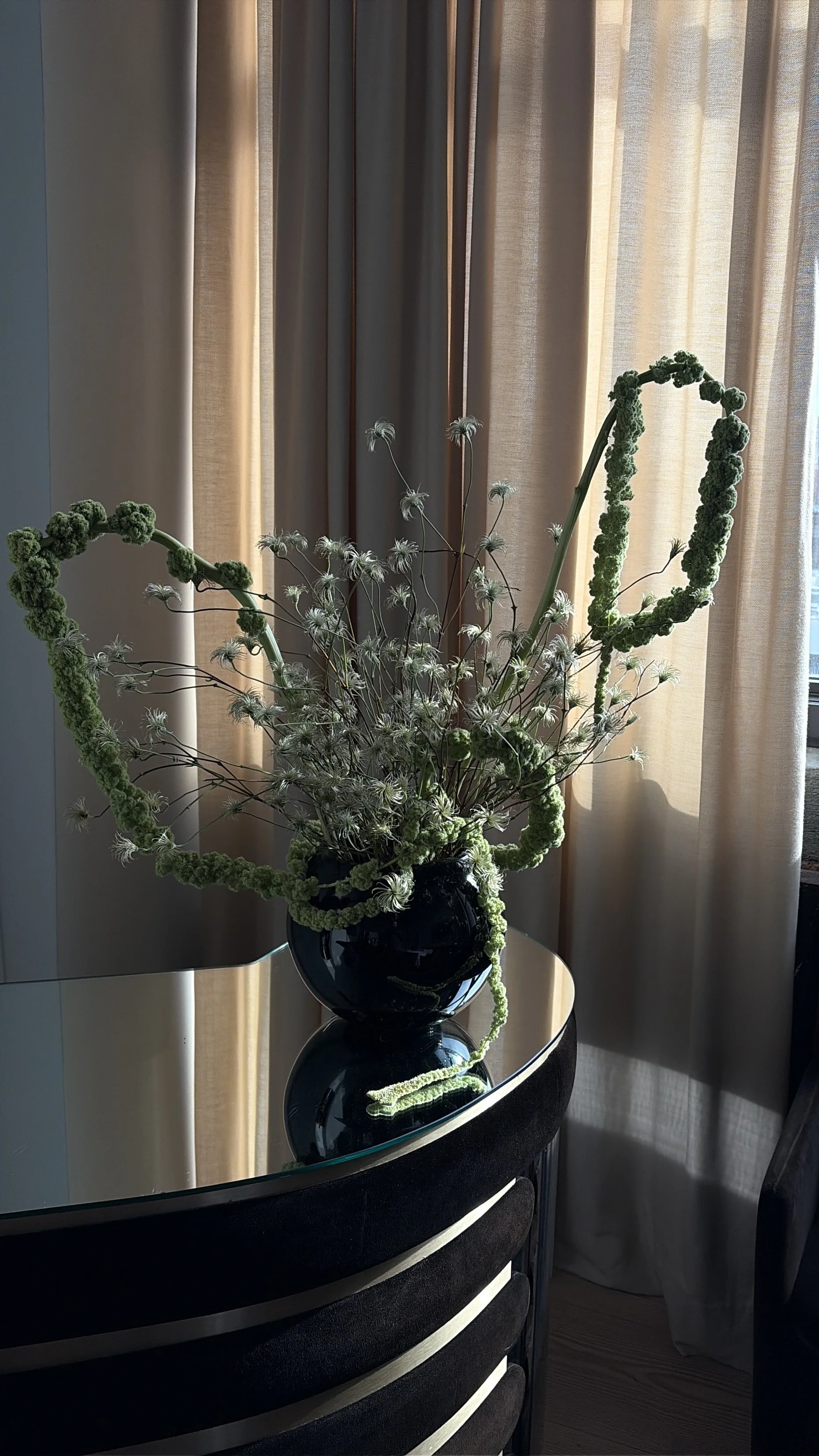 A flower arrangement in a black vase on a glass-top table, with green and white flowers and curly green accents, near beige curtains and a window.