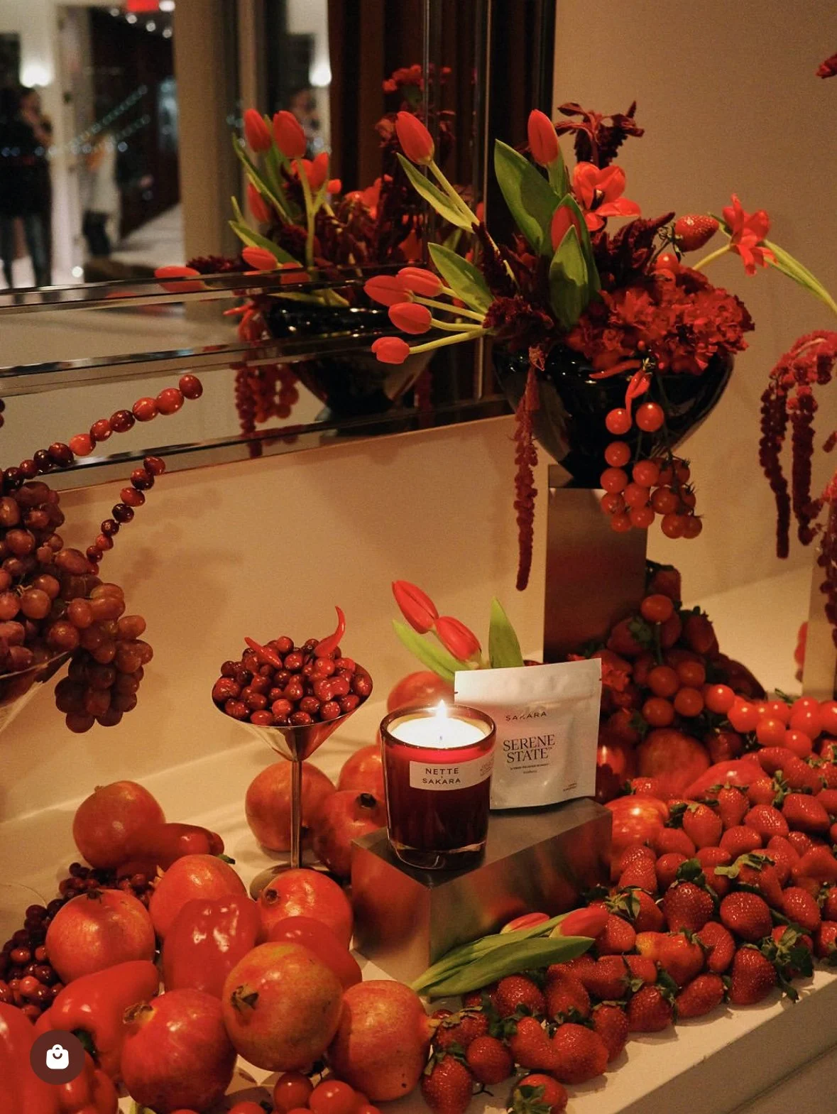 Arrangement of red flowers, strawberries, apples, and red berries with a candle, set on a table near a mirror.