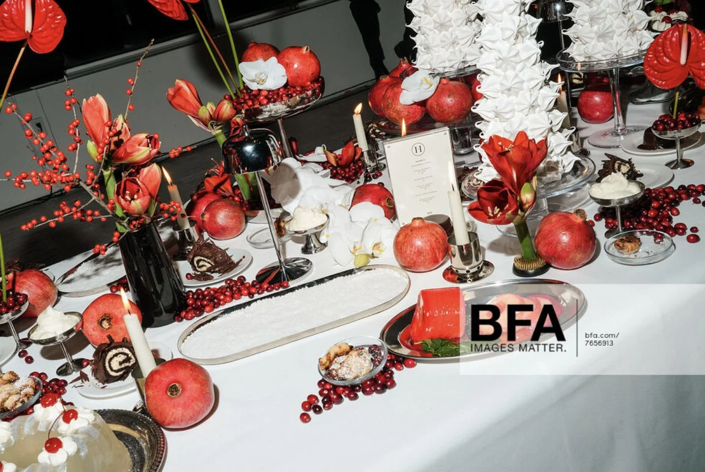 Table decorated with red and white flowers, pomegranates, apples, white candles, and assorted desserts including cakes and pastries.