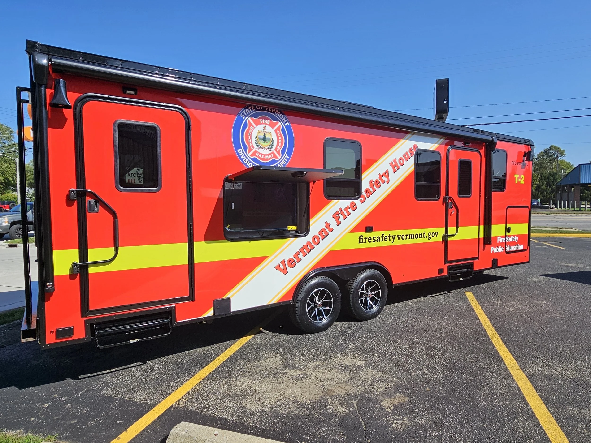 Red Vermont Fire Safety Education mobile trailer with yellow stripes, parked in a lot, featuring branding and a small window for public service.