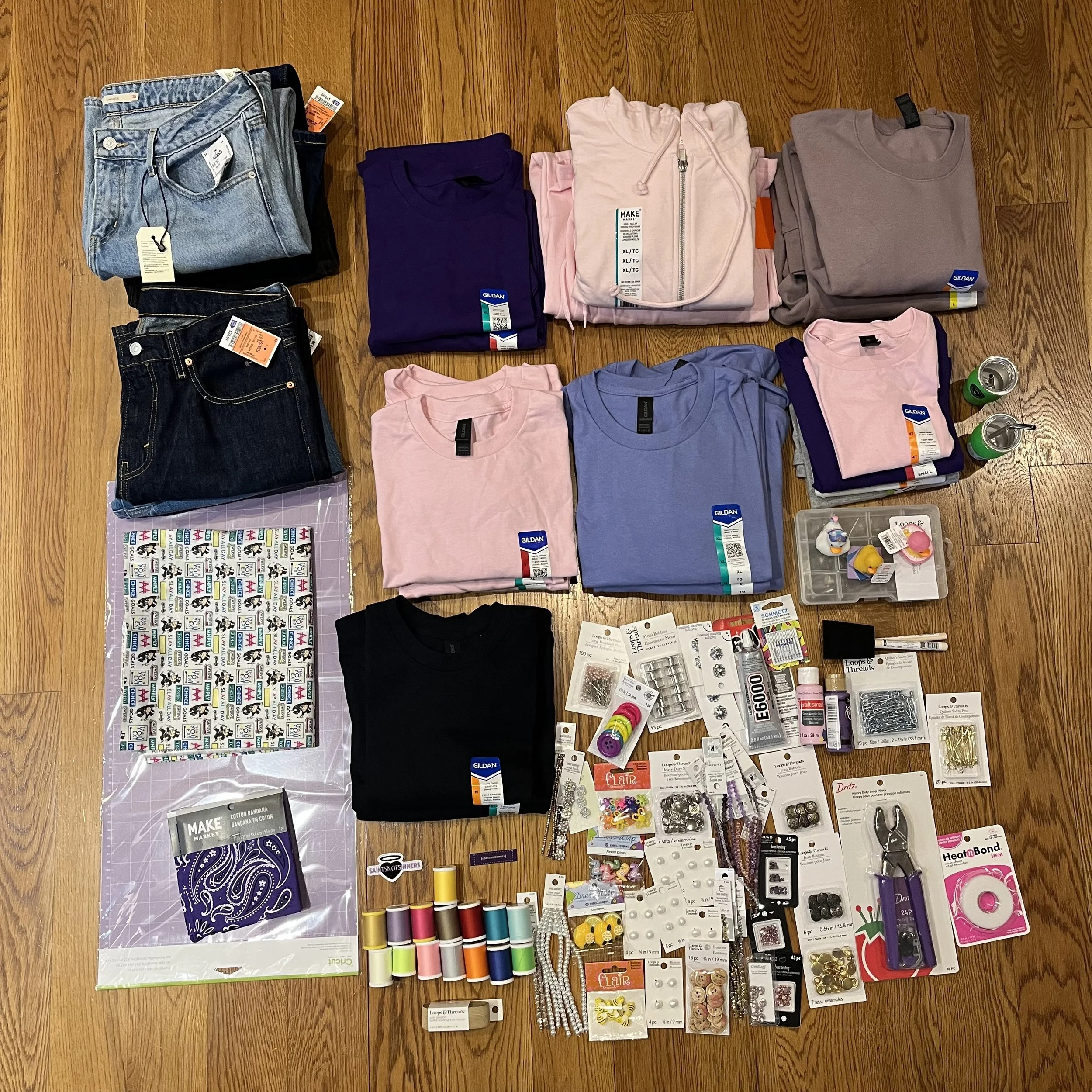 Clothes, sewing supplies, and accessories arranged on a wooden floor, including jeans, t-shirts, sweatshirts, sewing thread, ribbons, jewelry, pins, and other crafting items.