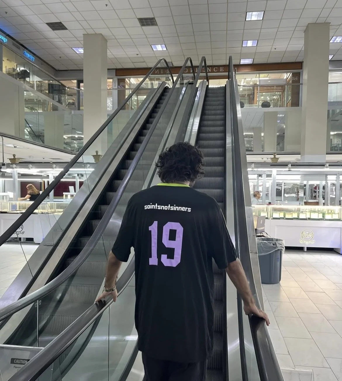Alam aka @grillzbyprince  wearing a black sports saintsnotsinners jersey with the number 19 using an escalator in the houston diamond district