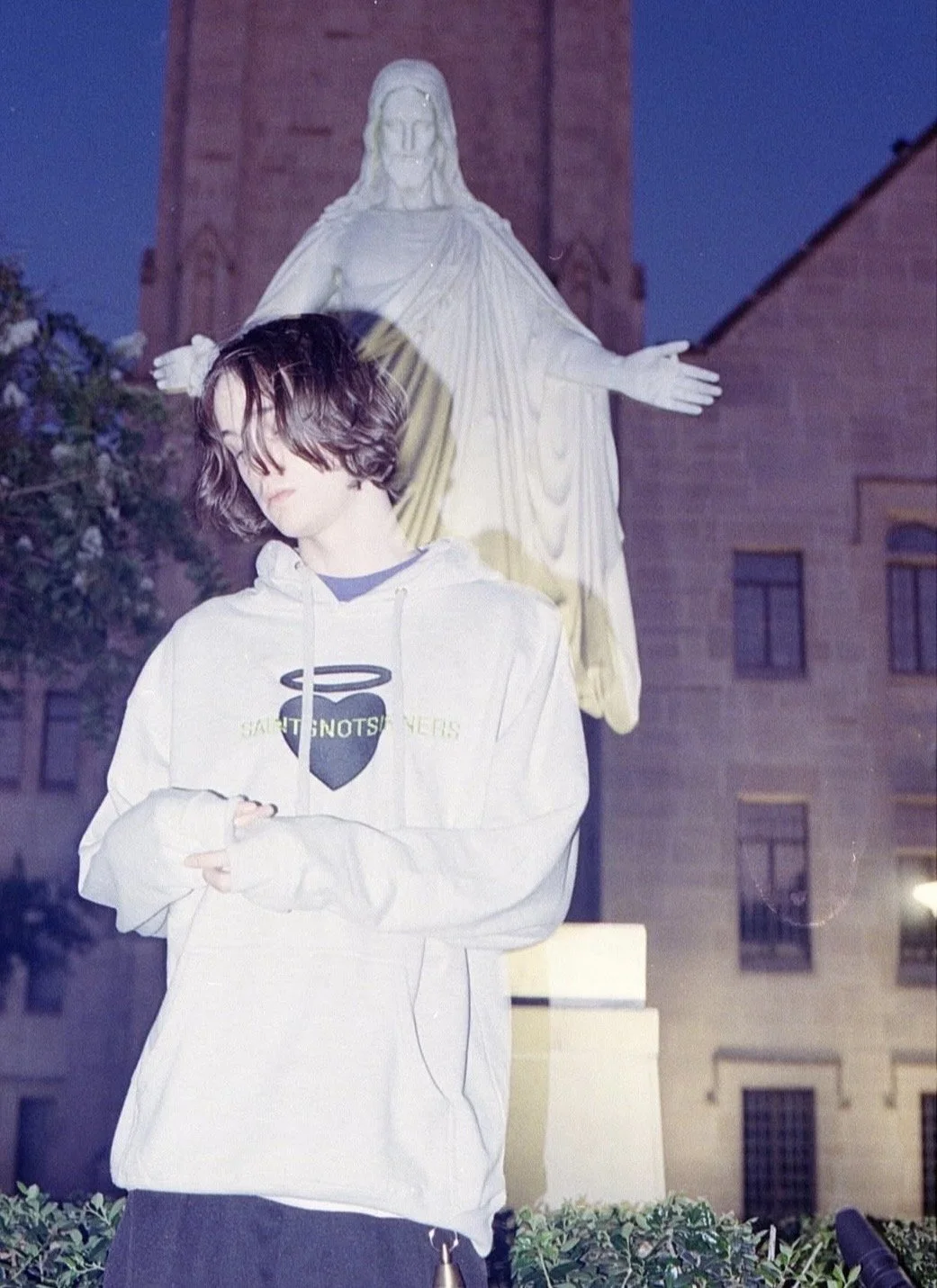 Young bro with dark, wavy hair standing outdoors at night in front of a large white statue of Jesus Christ, with a background of brick buildings and trees.