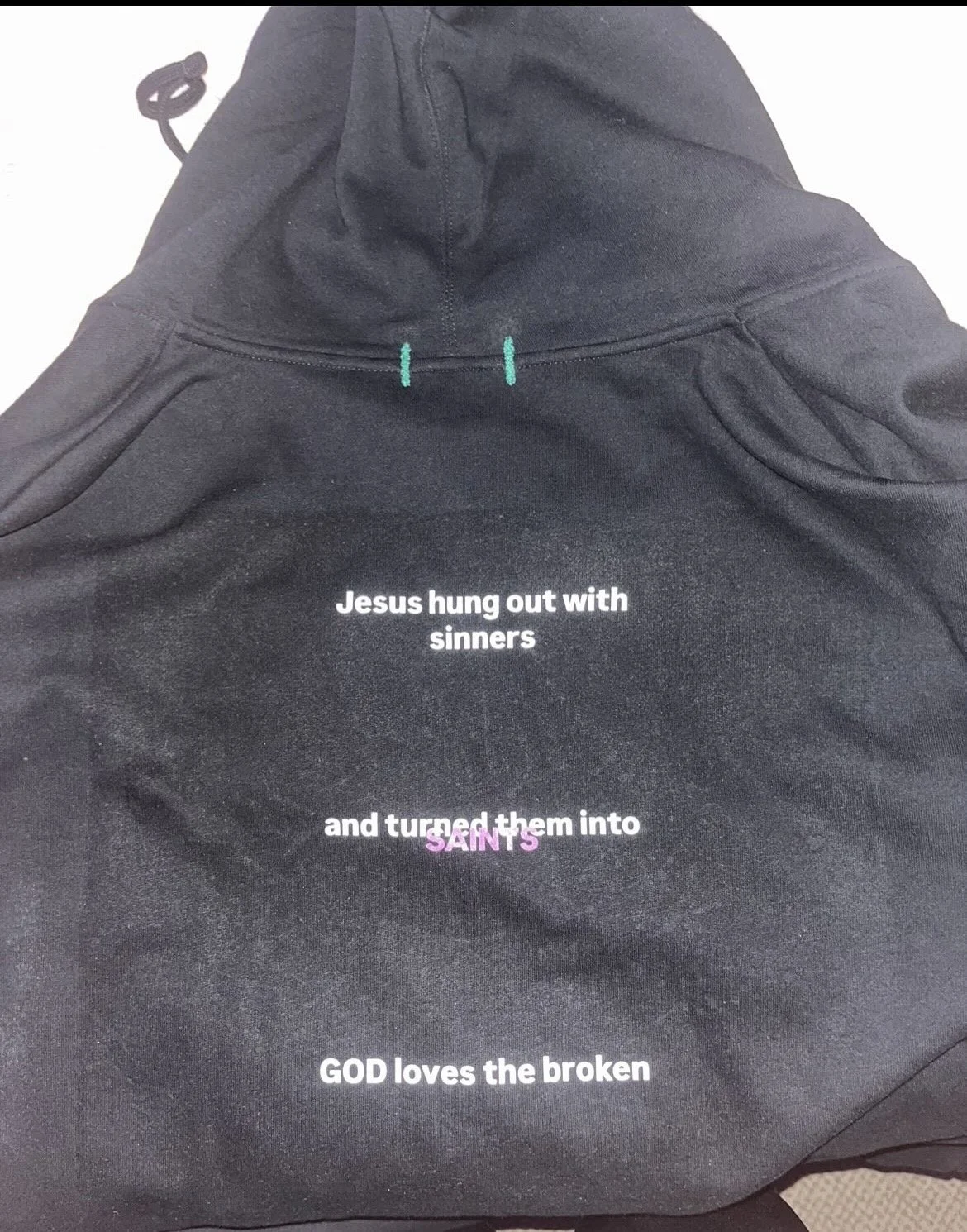 Black hoodie with white and purple text that reads, "Jesus hung out with sinners and turned them into saints. GOD loves the broken."
