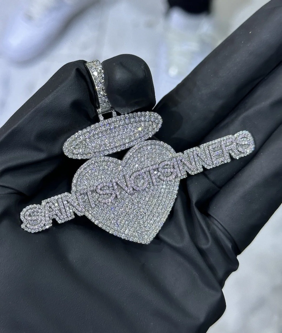 A gloved hand holding a large, diamond-encrusted necklace shaped like a heart and a halo with the words
 'SAINTSNOTSINNERS'  written across it, also decorated with diamonds.