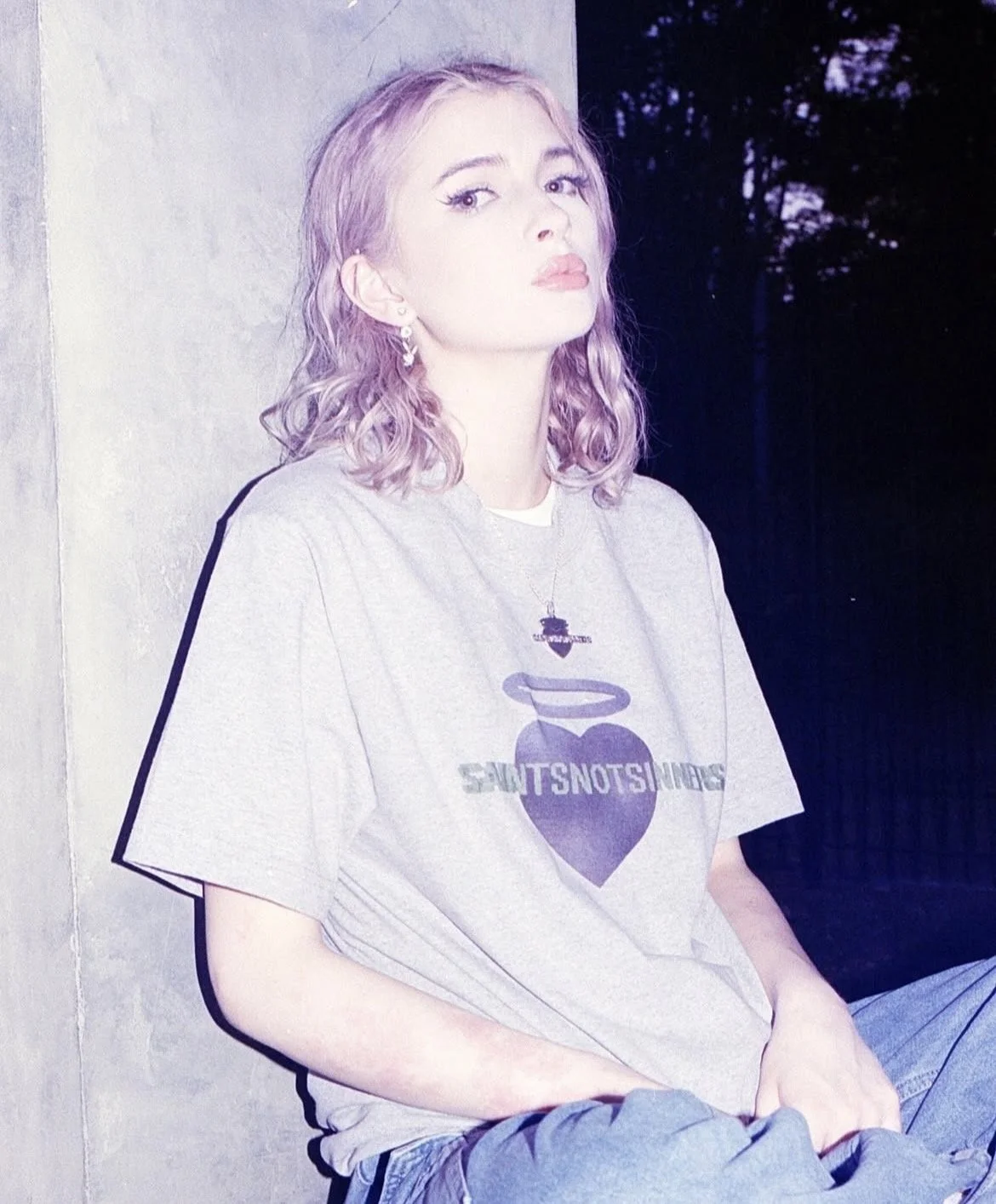 A young woman with shoulder-length wavy blonde hair sitting against a concrete wall at night, wearing a gray t-shirt with a purple heart and black text, earrings, and a necklace.