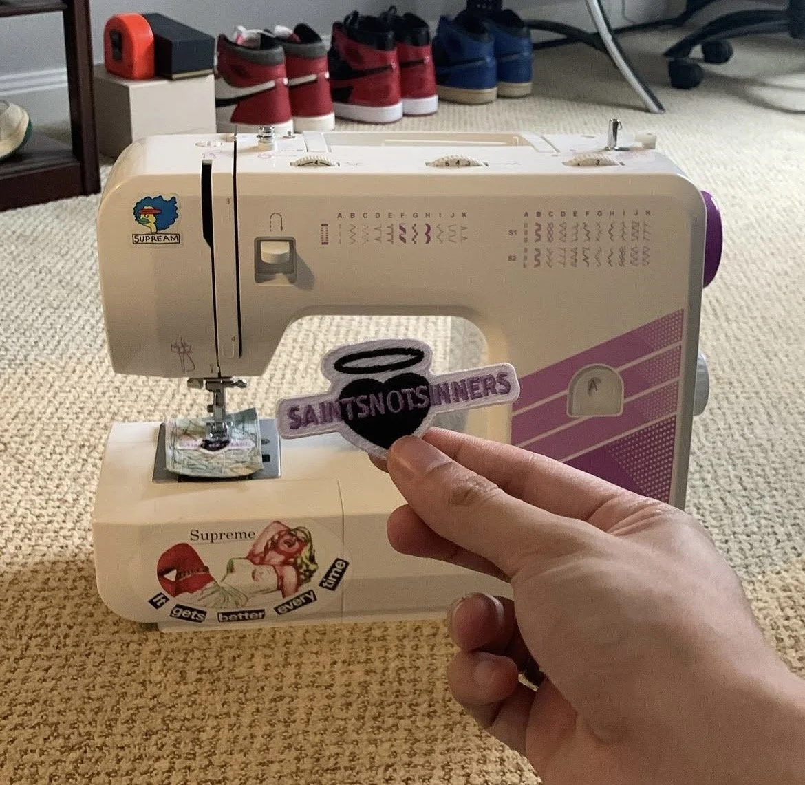 saint holding a patch that says 'SAINTSNOTSINNERS' in front of a sewing machine. The sewing machine is decorated with stickers and is placed on a beige carpeted floor, with several pairs of shoes in the background.