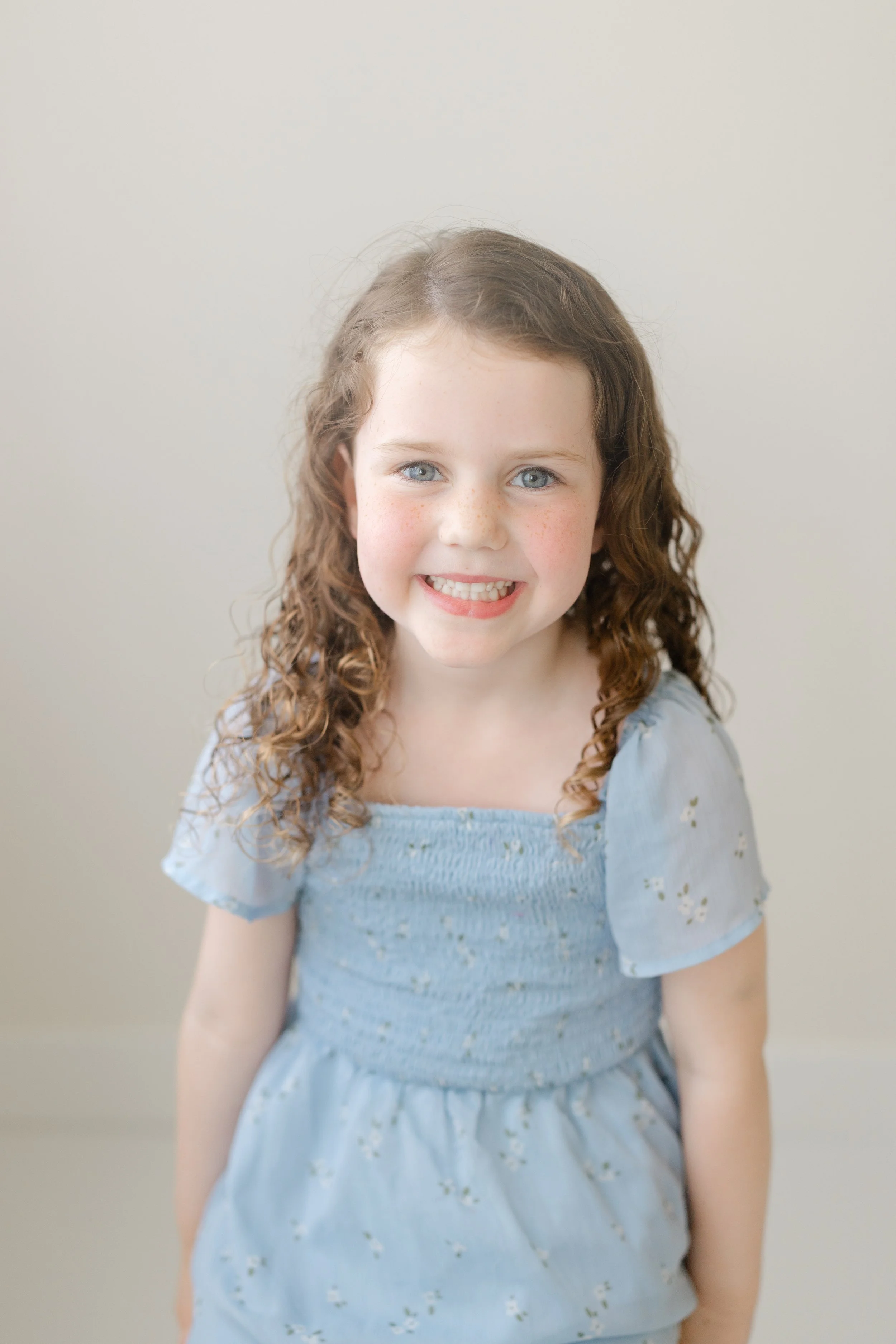 A young girl with curly light brown hair, blue eyes, and freckles, smiling and wearing a light blue dress with puffed sleeves and floral embroidery, standing against a plain beige background.