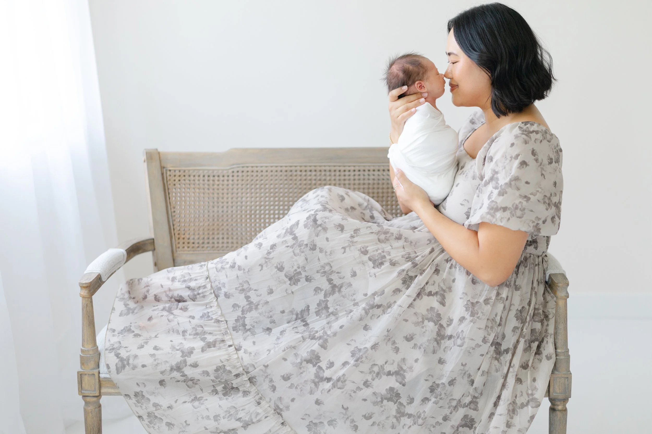 A woman with short black hair holding a baby close to her face in a softly-lit room with a white wall and light-colored furniture.