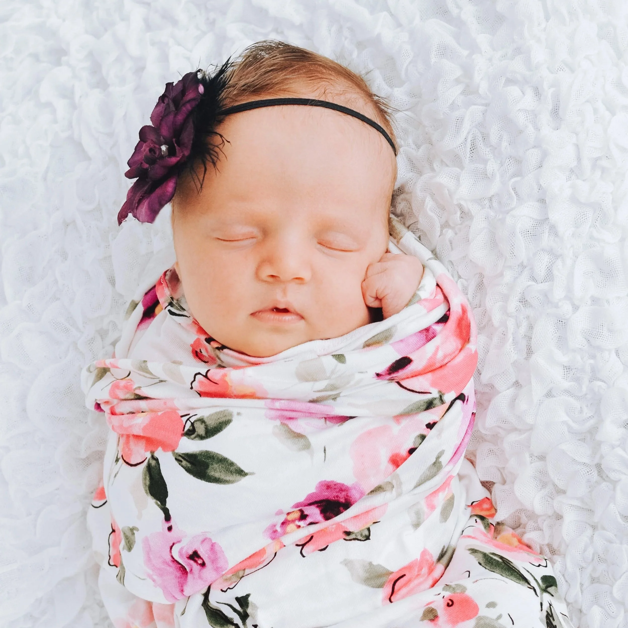 Close-up of a sleeping baby with a purple flower headband, lying on a white textured blanket.