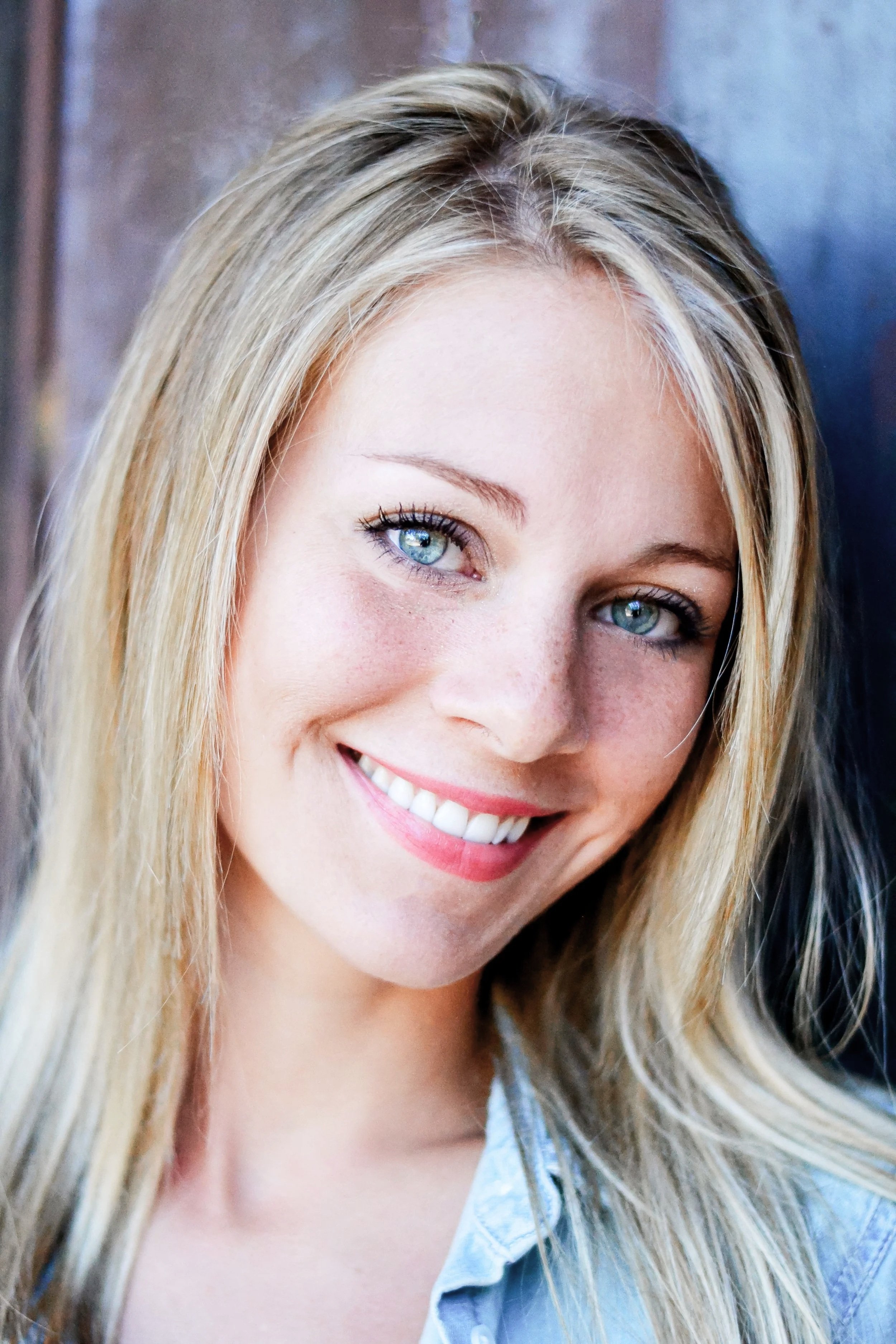 Close-up of a woman with blonde hair, blue eyes, and a wide smile, wearing a light blue shirt.