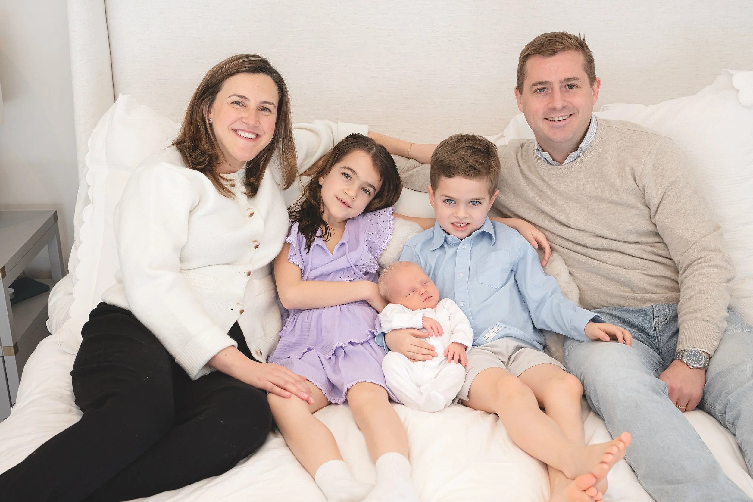 A happy family sitting together on a bed in a well-lit room, including two adults, two children, and a newborn baby.