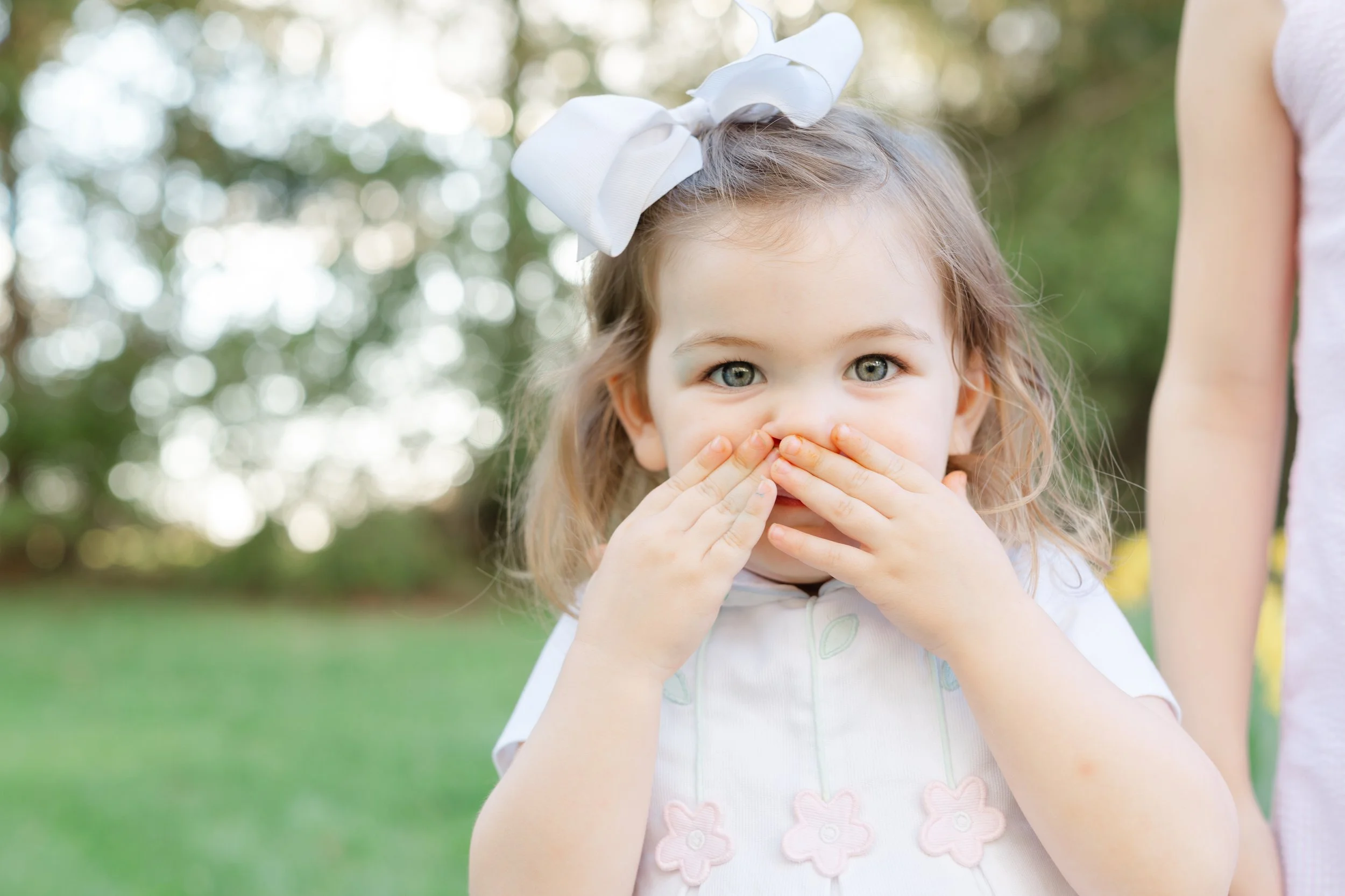 A young girl with blonde hair and blue eyes, wearing a white dress with pink flower designs, is covering her nose and mouth with her hands. She has a large white bow in her hair, and her expression appears playful. The background shows trees with blu