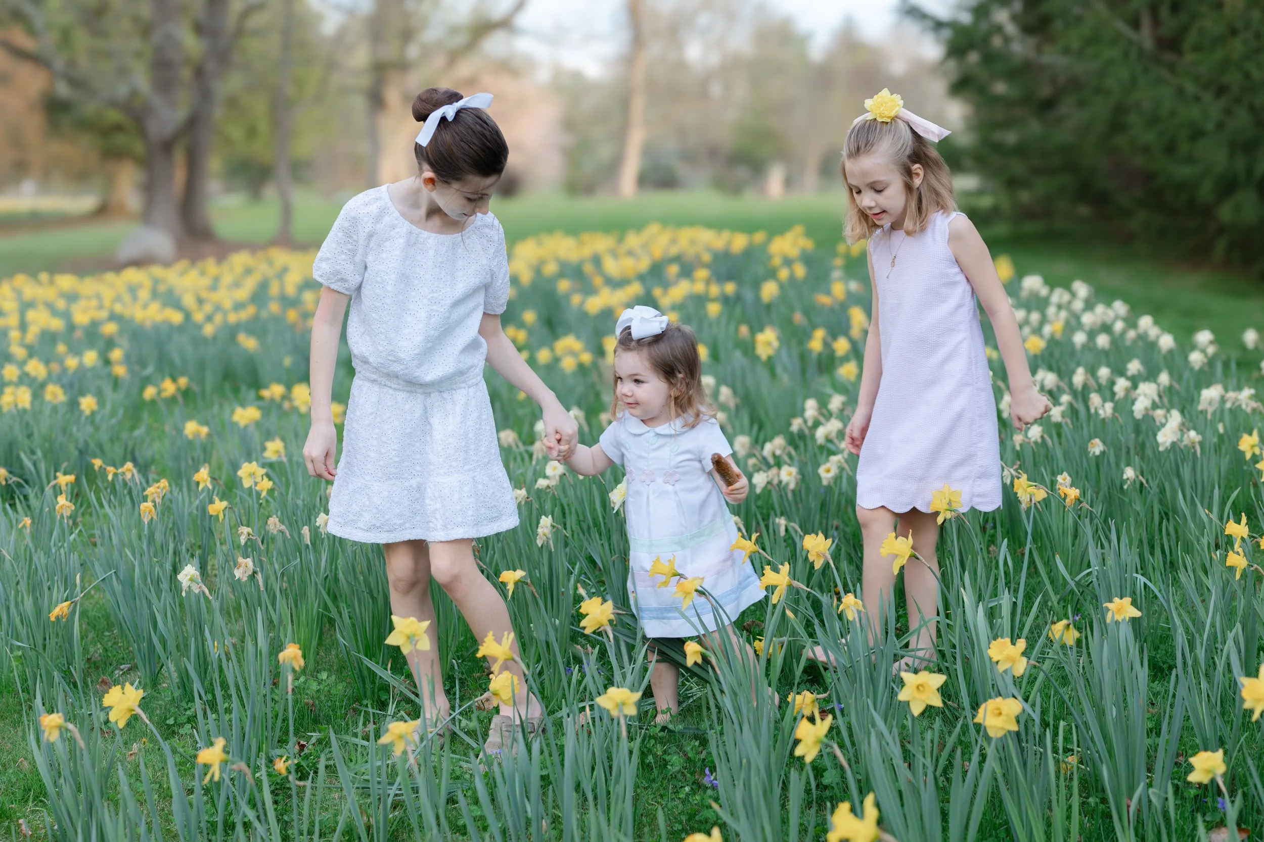 Three young girls in white dresses walking through a field of yellow and white flowers, holding hands, with trees in the background.