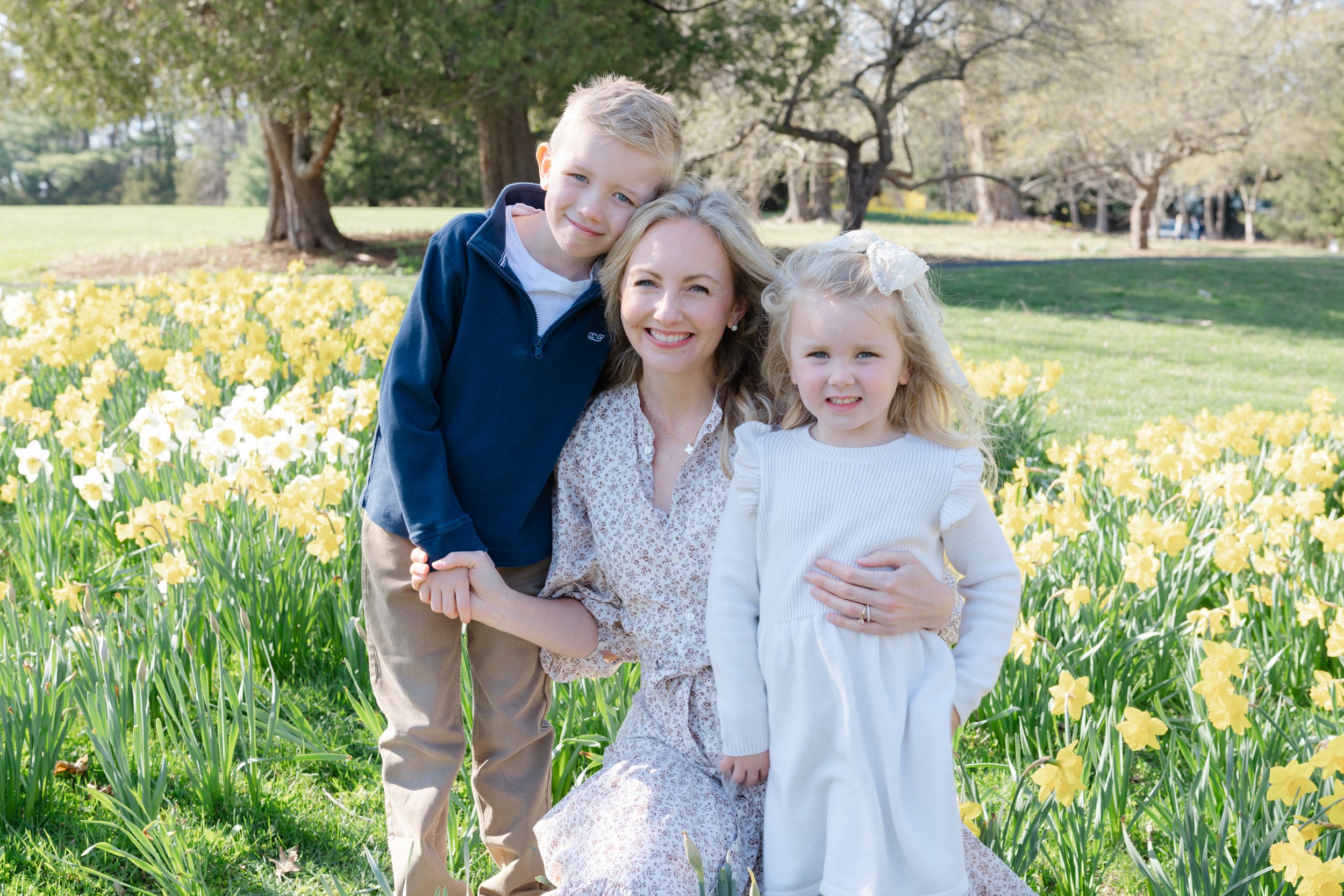 A woman with two children, a boy and a girl, posing in a field of yellow flowers with trees in the background.
