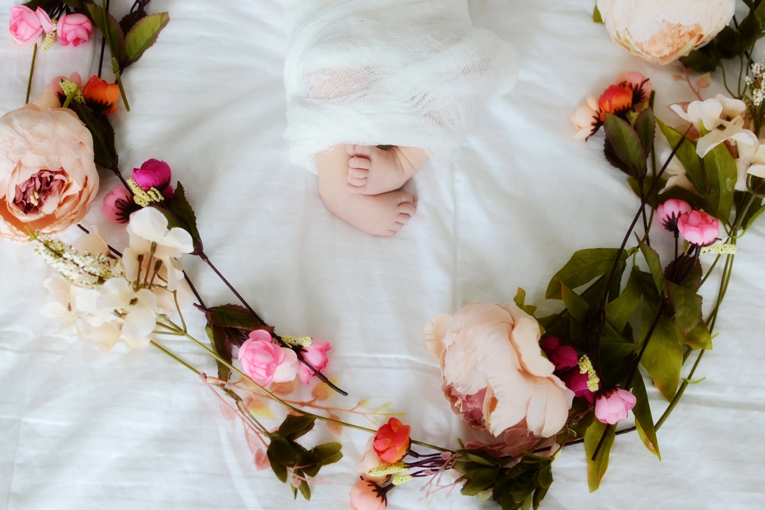 A newborn baby's feet covered with a white cloth, surrounded by pink, white, and peach-colored flowers arranged in a circular pattern on a white surface.