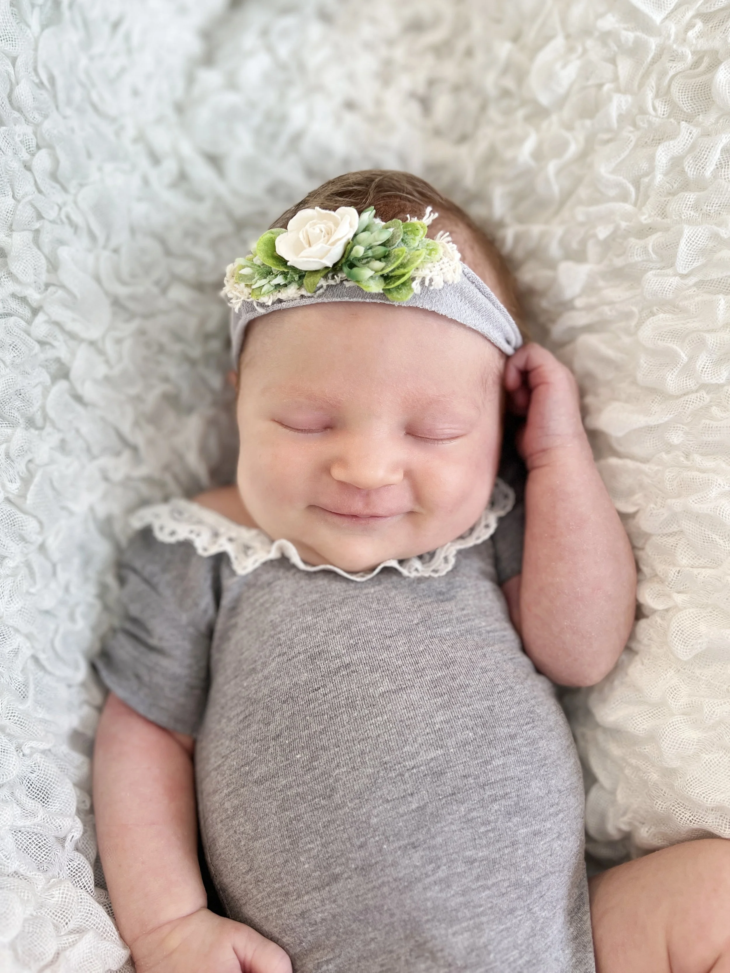 Close-up of a smiling newborn baby sleeping on a white textured blanket, wearing a gray outfit and a floral headband with white and green flowers.