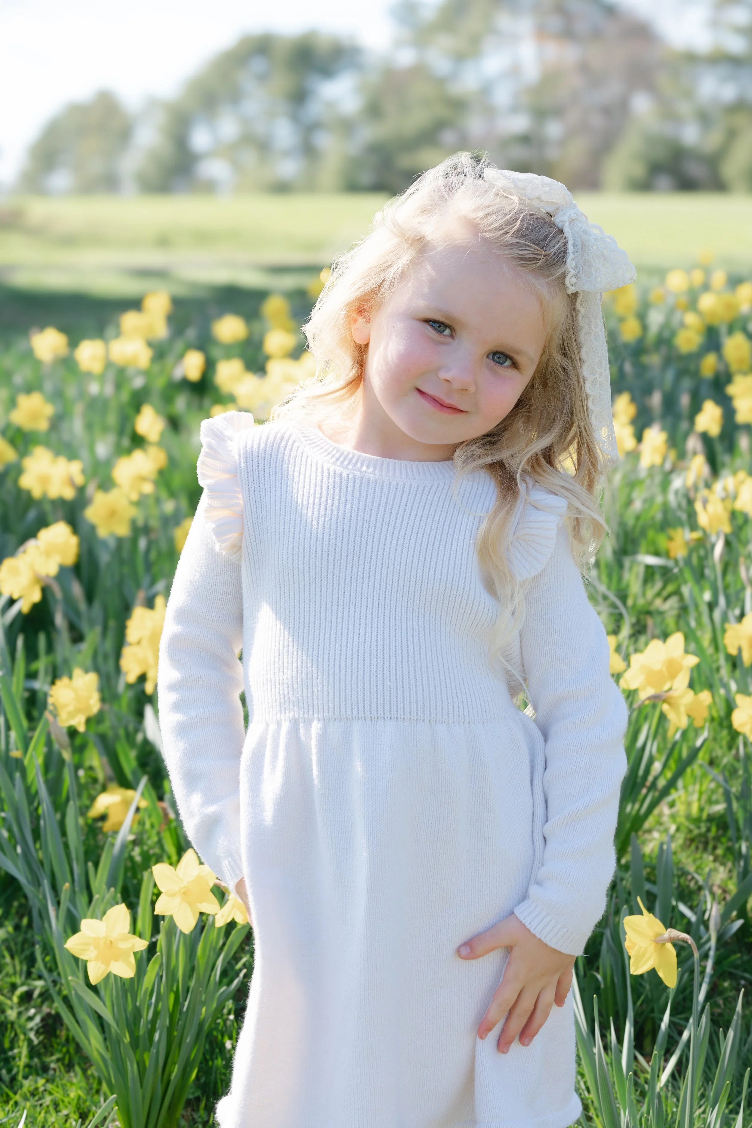 A young girl with blond hair and blue eyes, wearing a white dress and a lace bow, standing in a field of yellow flowers on a sunny day.