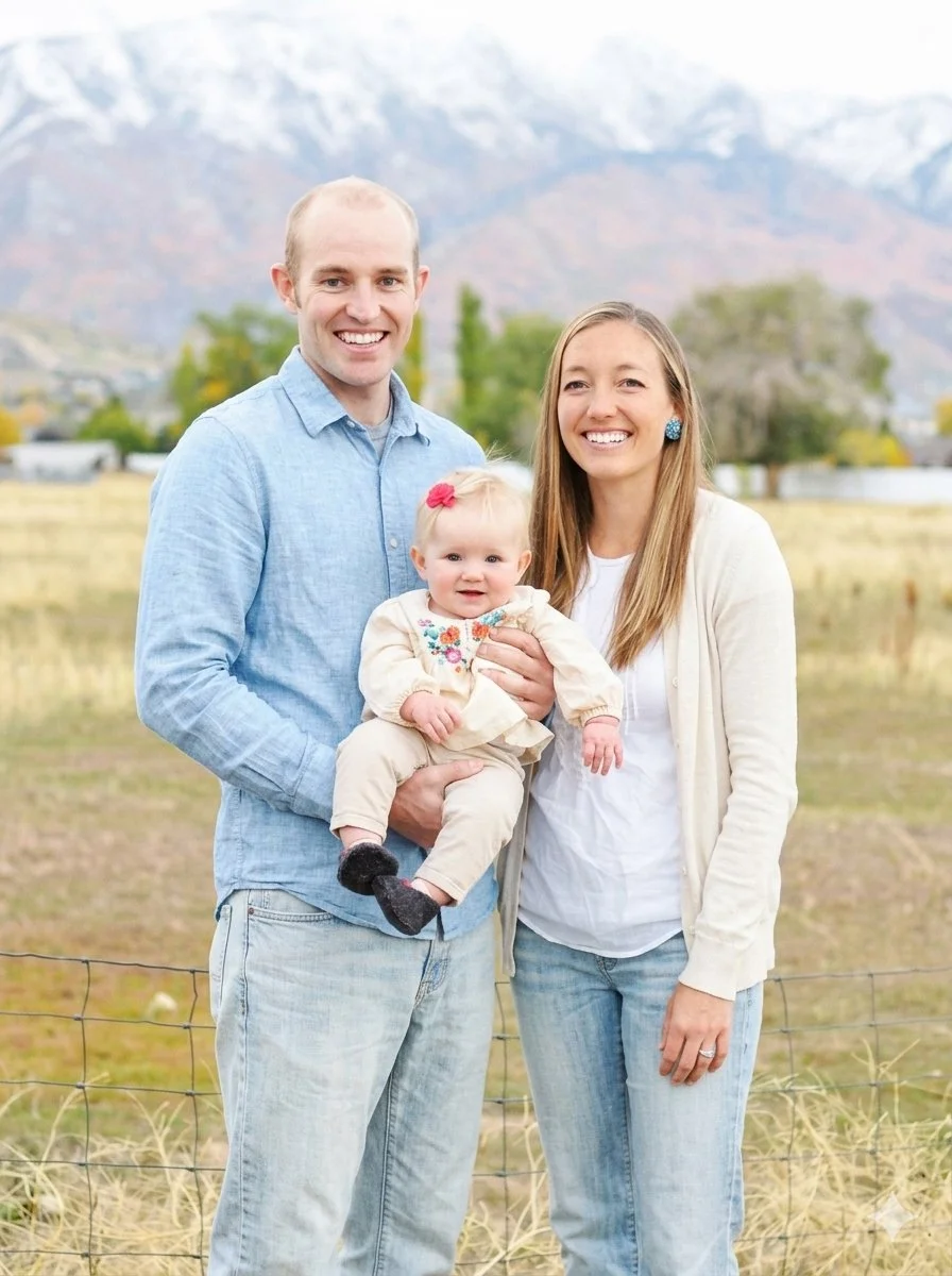 A smiling family of three stands outdoors in a field with mountains in the background. The man holds a toddler girl, and the woman stands beside him.