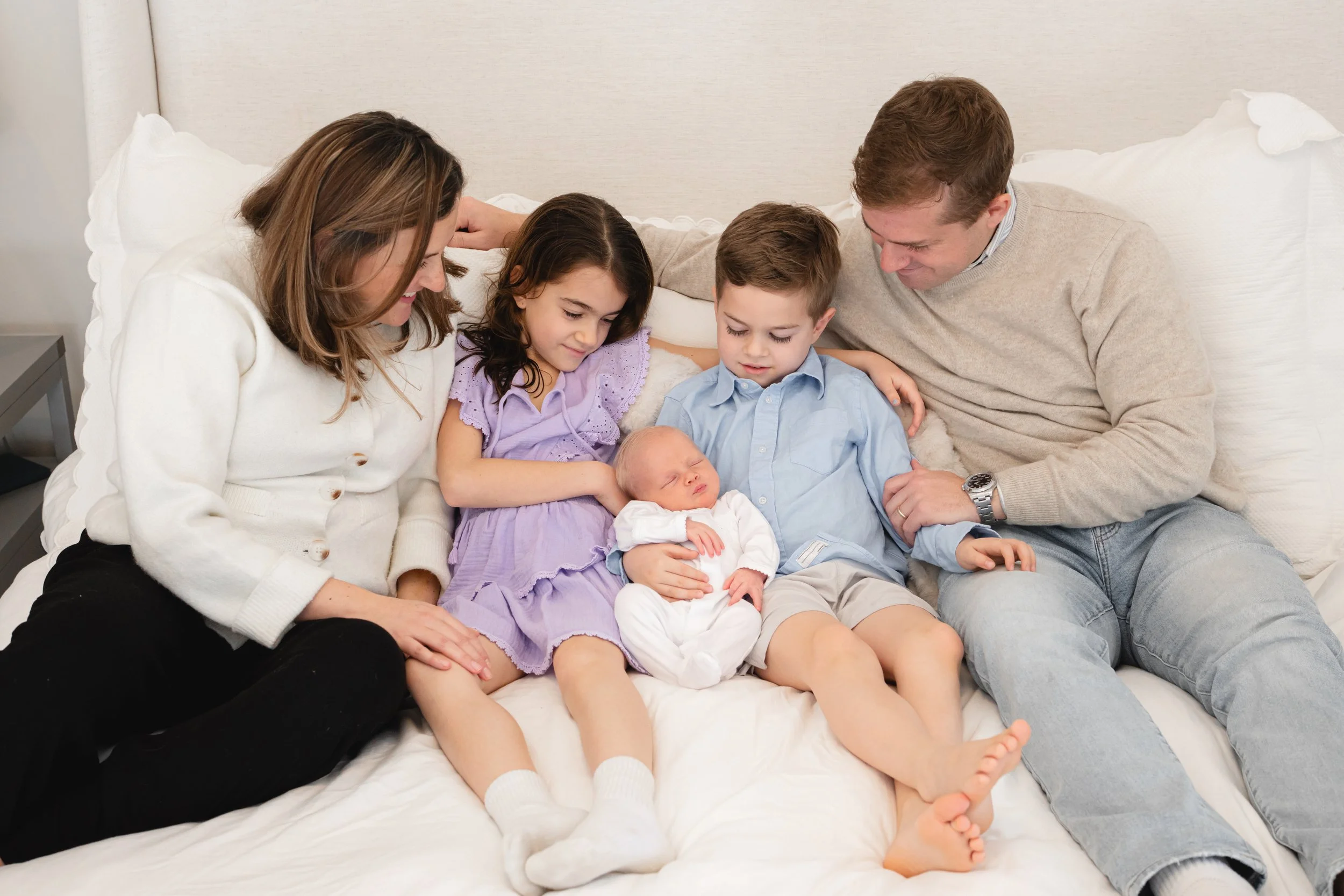 A multi-generational family sitting on a bed, gathered around a newborn baby. The family includes two young children, parents, and an older woman, all smiling and looking affectionately at the baby.