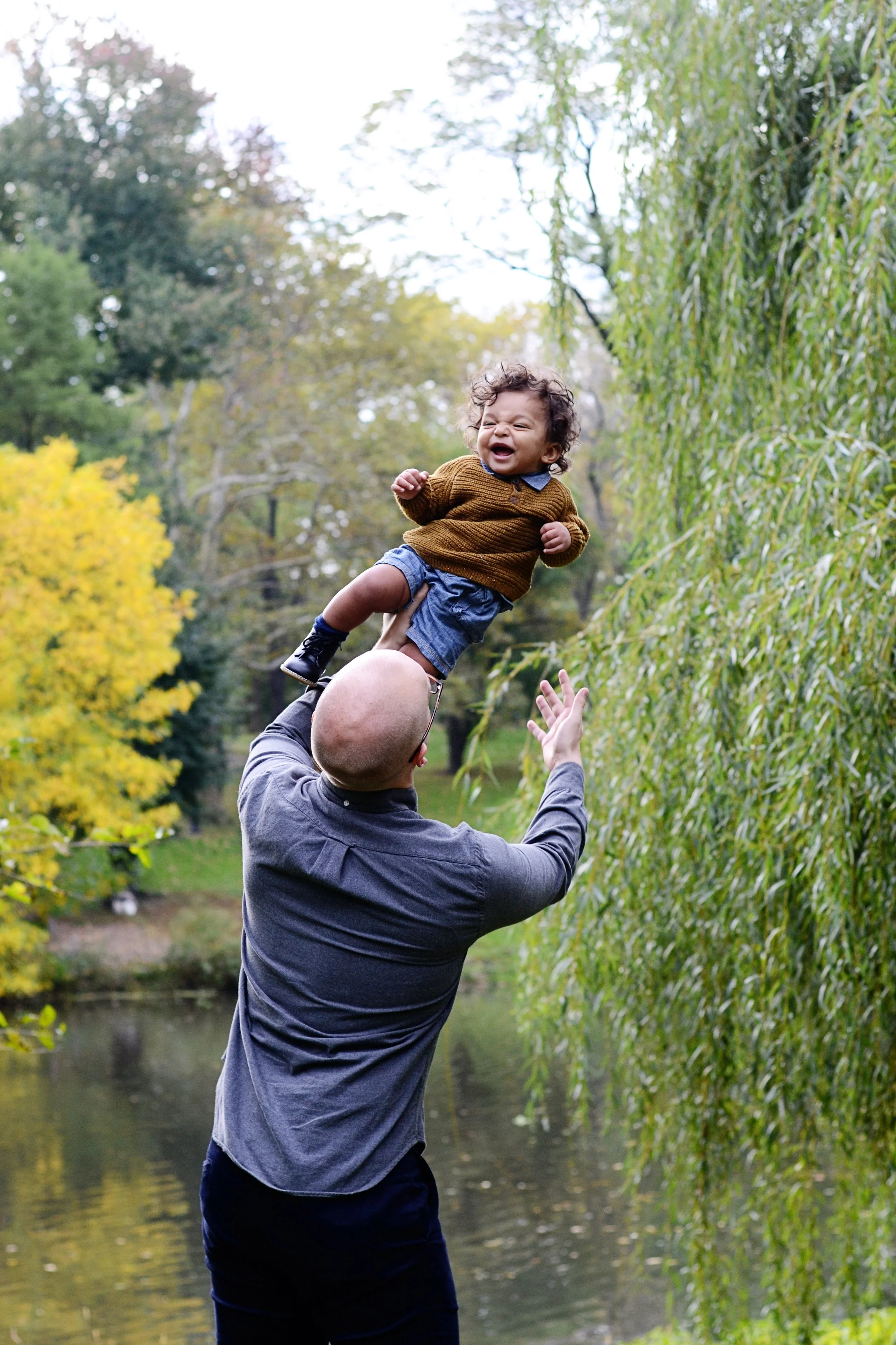 A man is holding a child up in the air outdoors near a body of water, surrounded by trees with green and yellow leaves, during daytime.