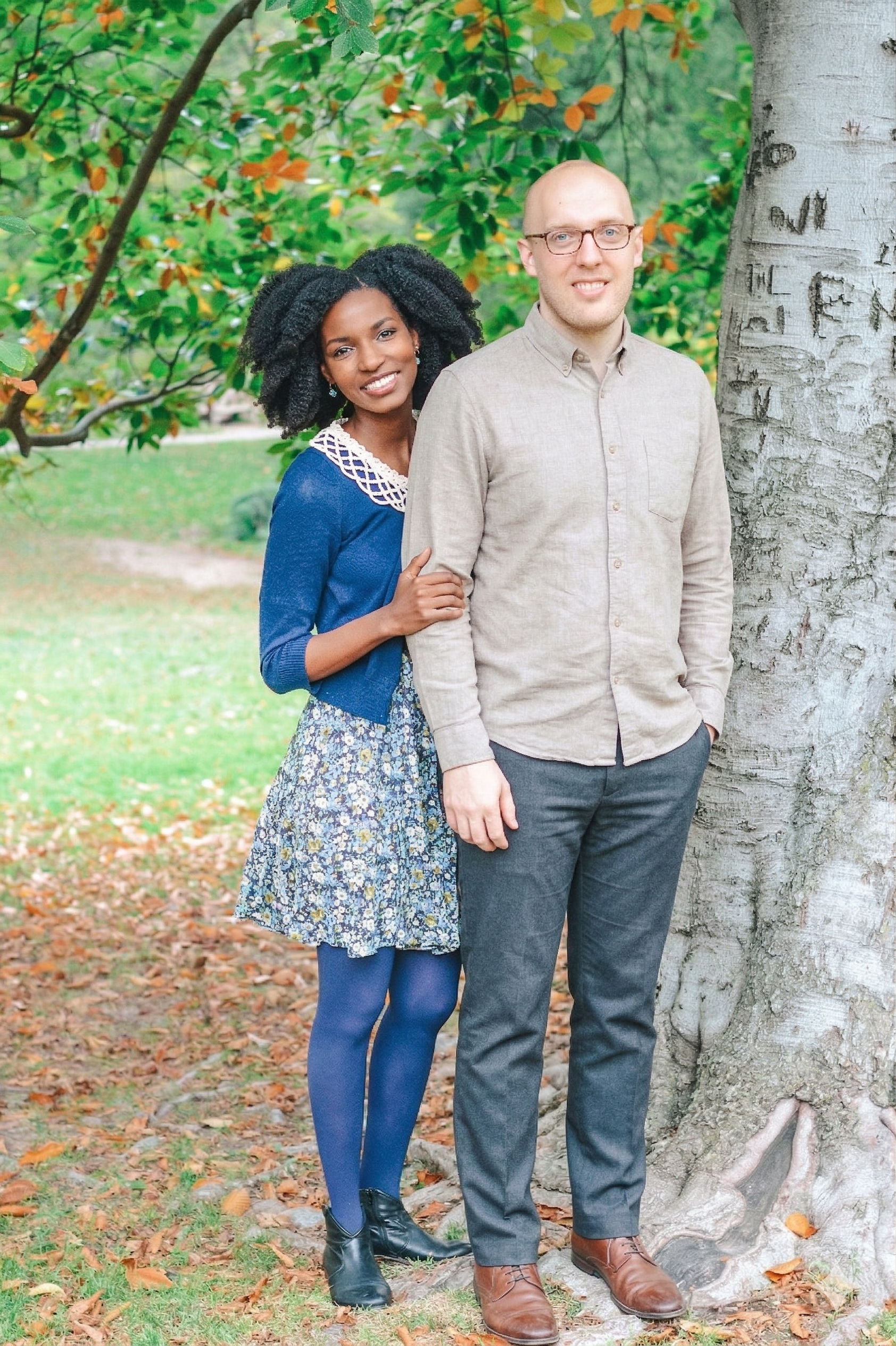 A couple standing outdoors near a tree in a park. The woman has curly black hair, is wearing a blue cardigan, a floral skirt, blue tights, and black ankle boots. The man is bald, wearing glasses, a beige button-down shirt, dark gray pants, and brown 