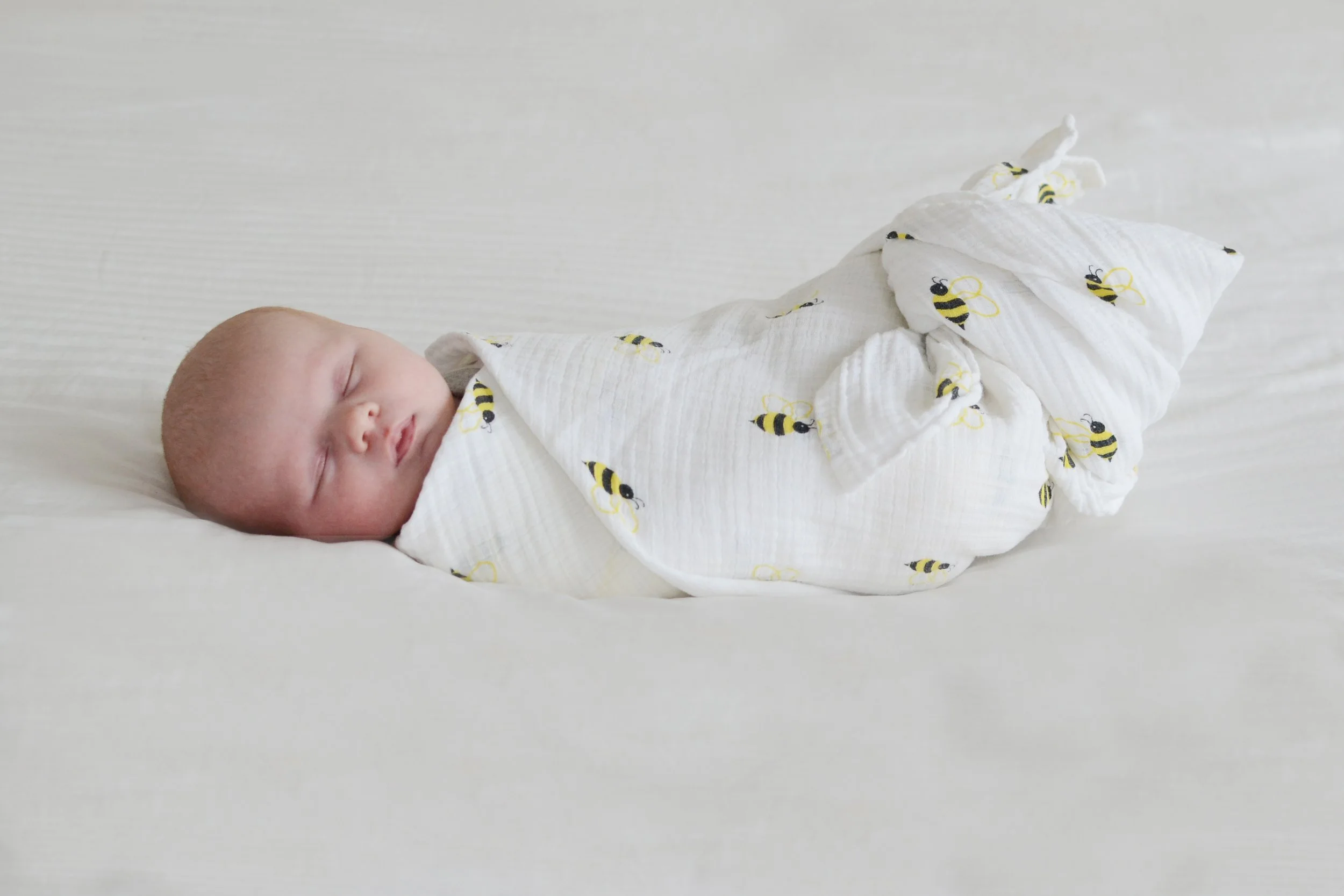 A sleeping baby wrapped in a white blanket with yellow and black bee patterns, lying on a white surface.