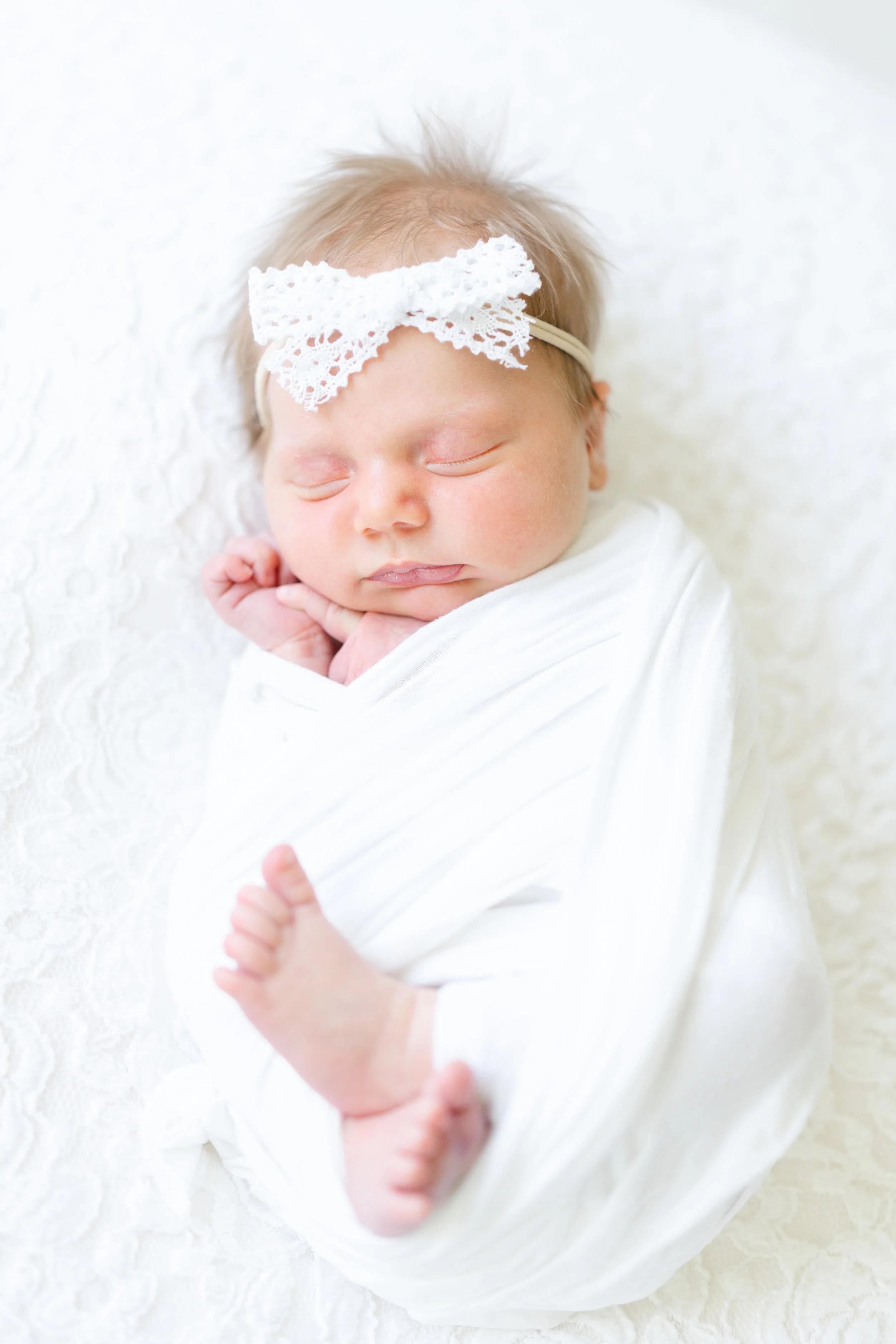A sleeping newborn baby wrapped in a white blanket with a lace headband on a white textured background.