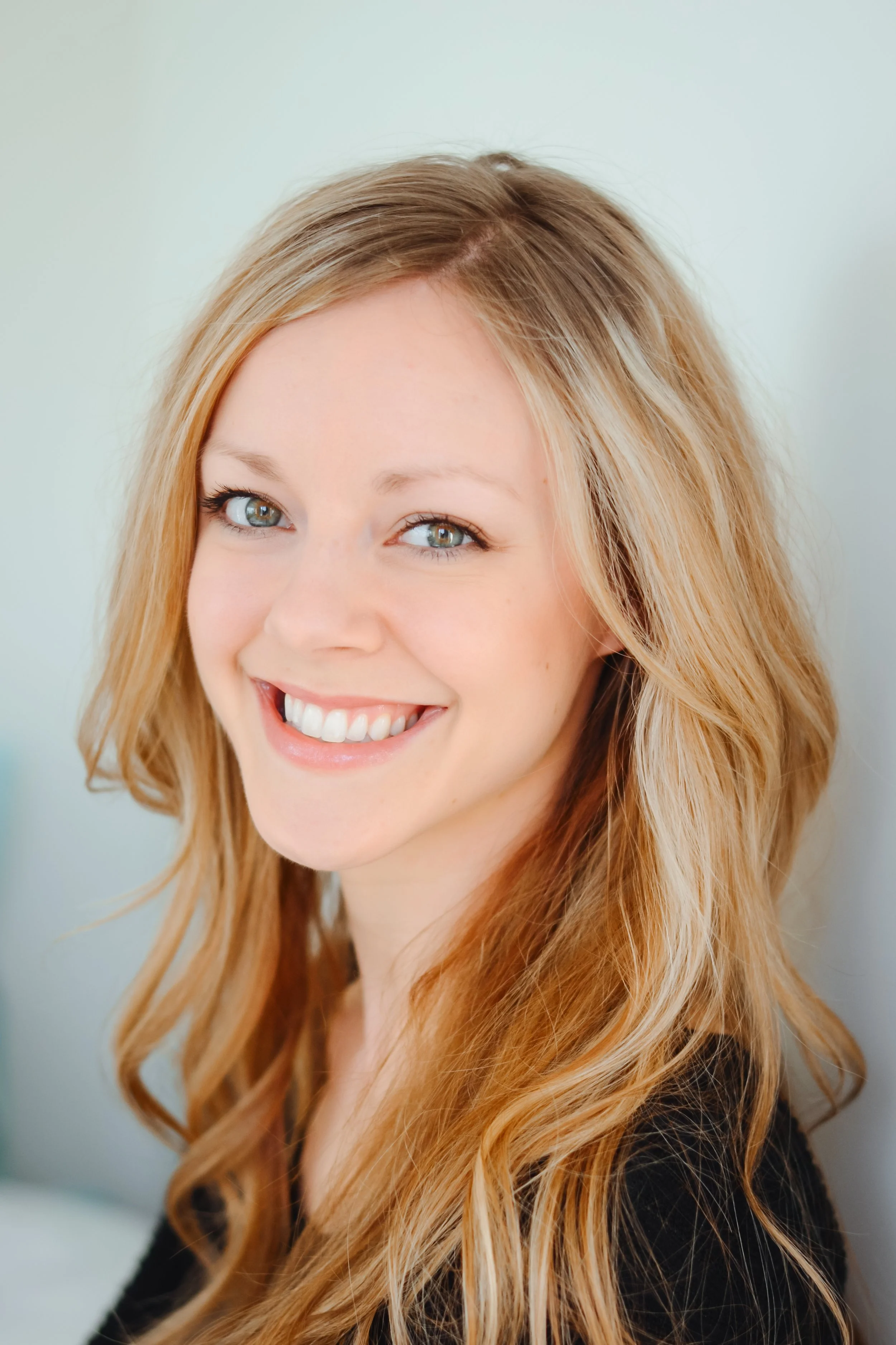 Close-up portrait of a smiling woman with long, wavy, strawberry blonde hair and blue eyes, wearing a black top.