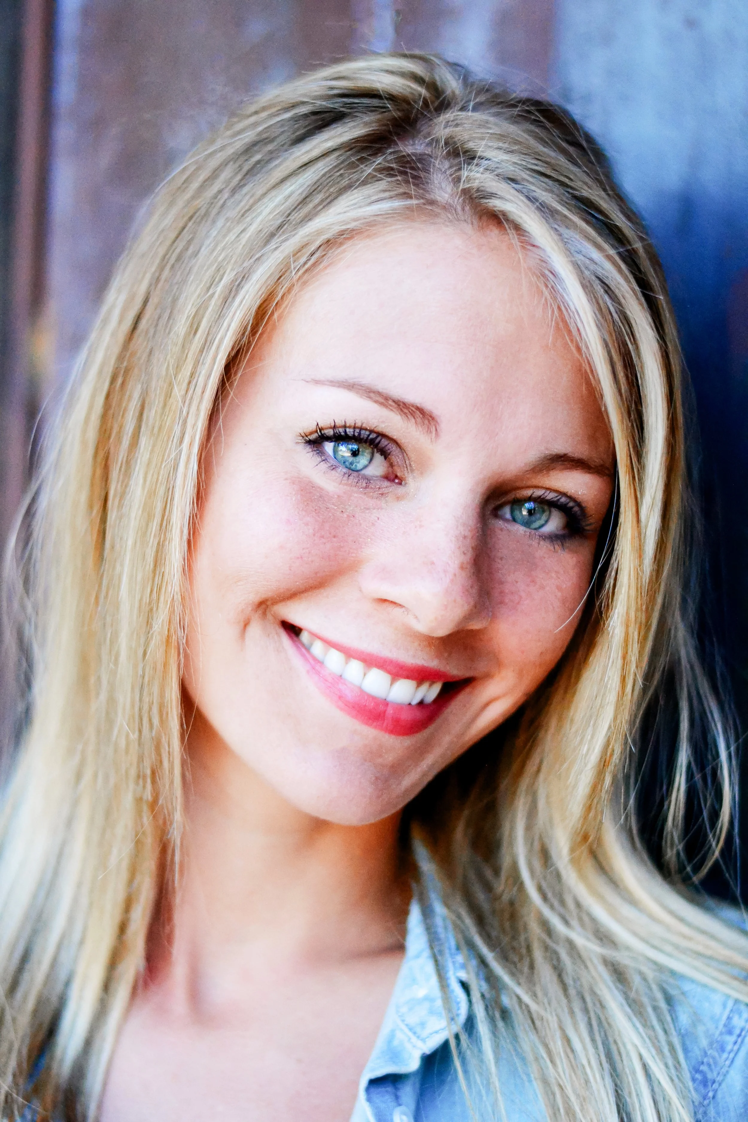 Close-up of a smiling woman with long blonde hair, blue eyes, and wearing a light blue collared shirt.