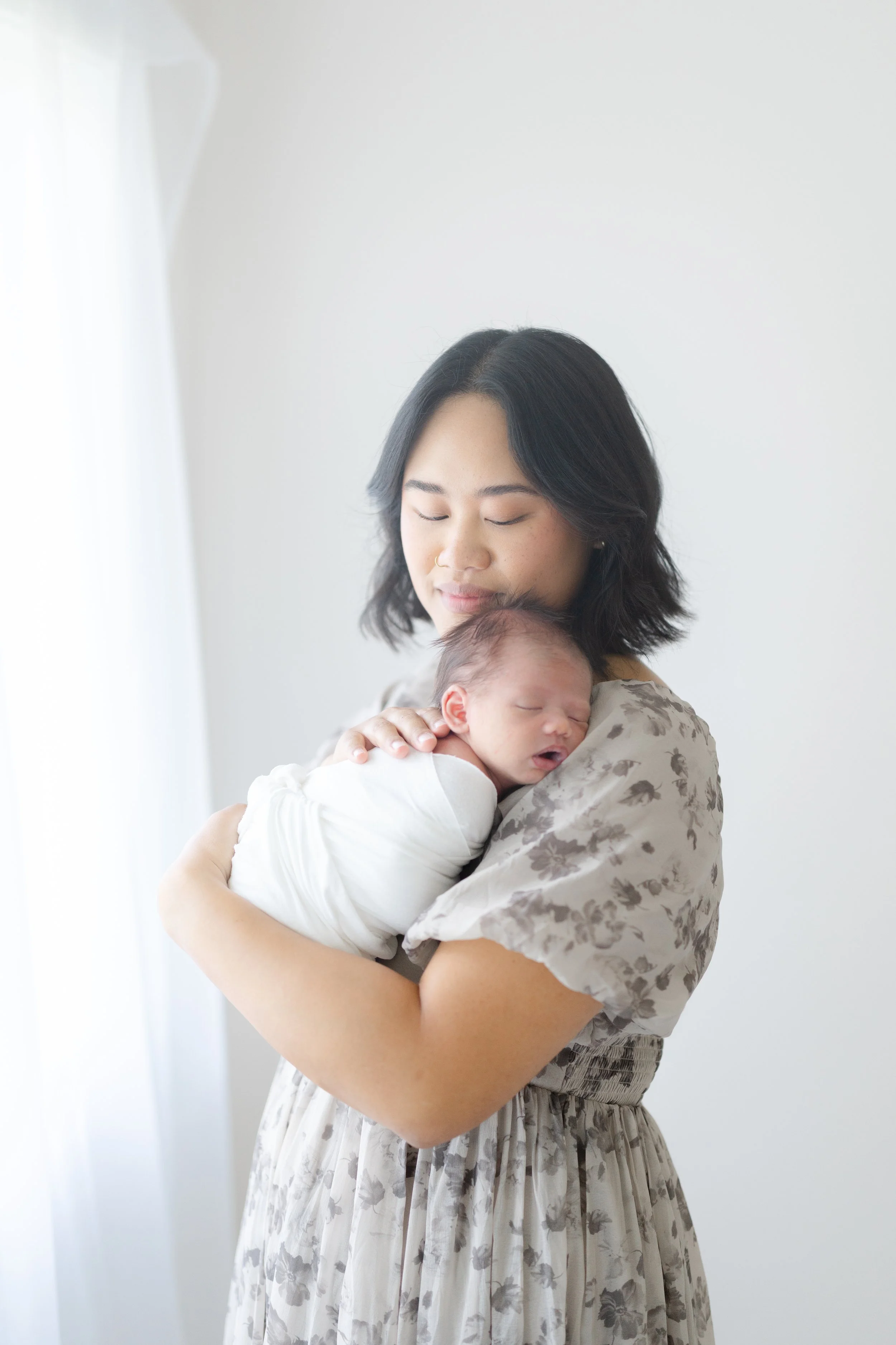 A woman with dark hair holding a sleeping newborn baby in her arms inside a bright room with white walls and curtains. Connecticut Photographer. Connecticut Newborn Photographer. Family Photographer in Connecticut. CT Newborn Photographer.