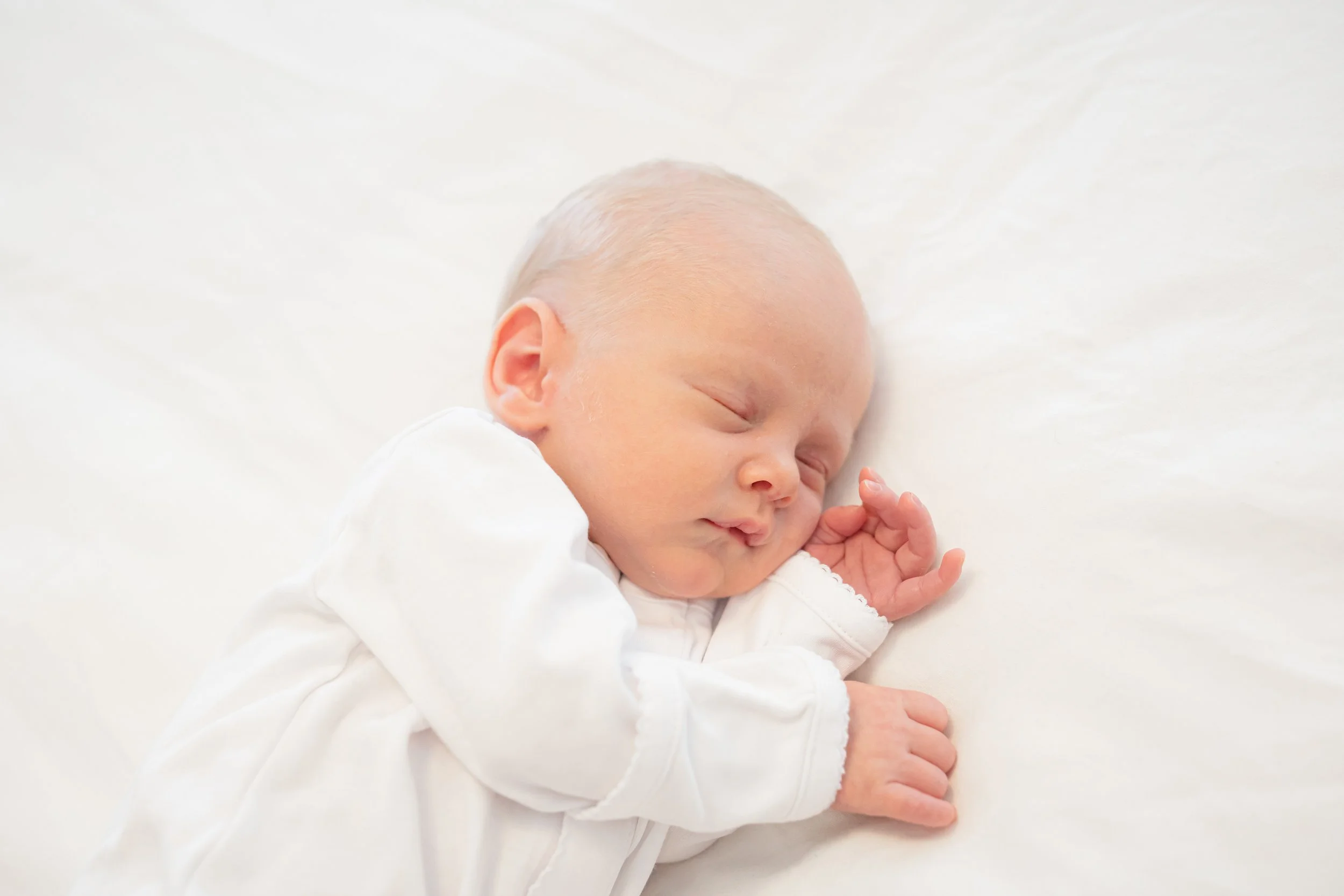 A baby with light skin and short blond hair is peacefully sleeping on a white blanket, dressed in a white outfit, with hands gently clasped near their face.