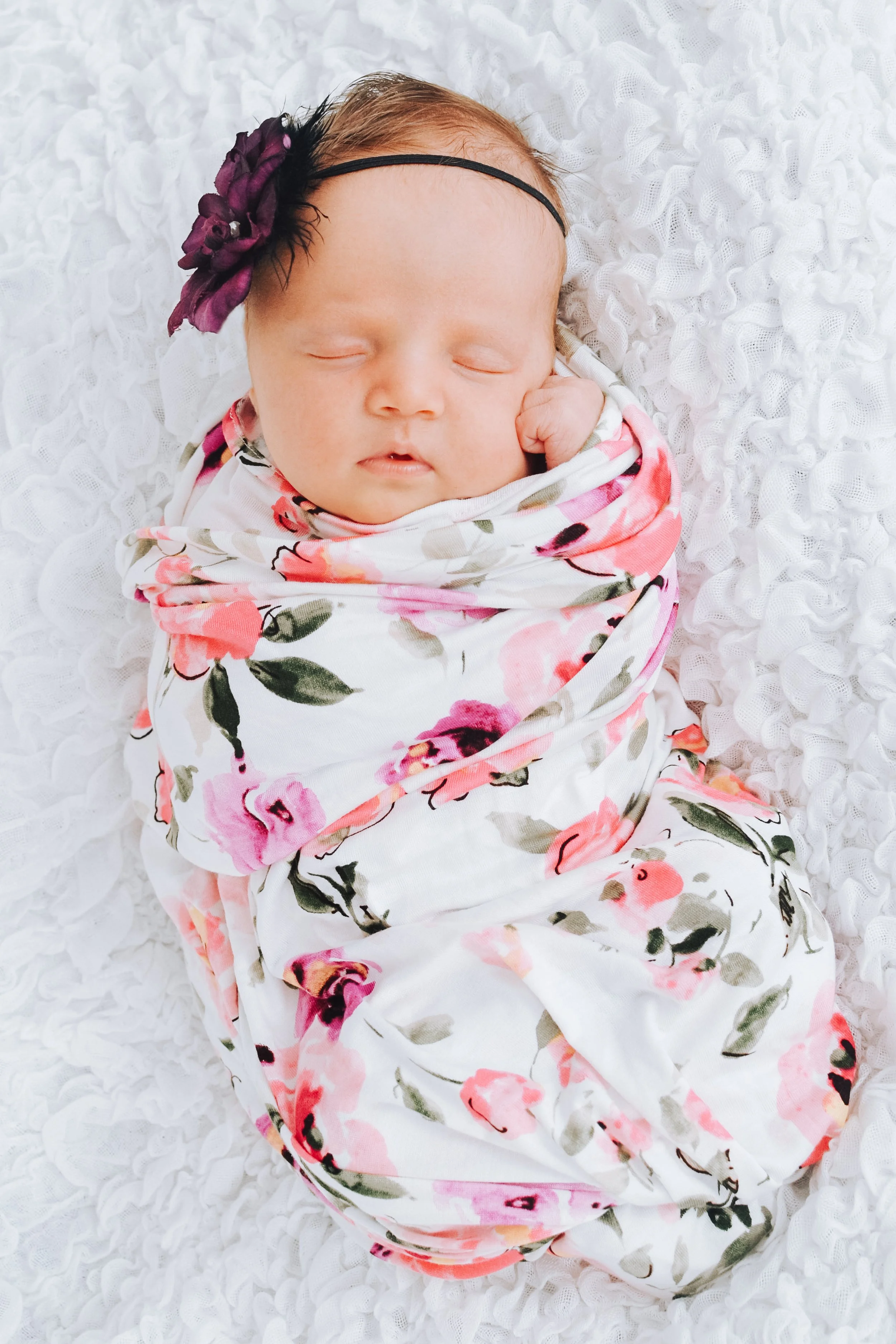 A sleeping baby girl with a purple flower headband, wrapped in a floral blanket, lying on a white textured blanket.
