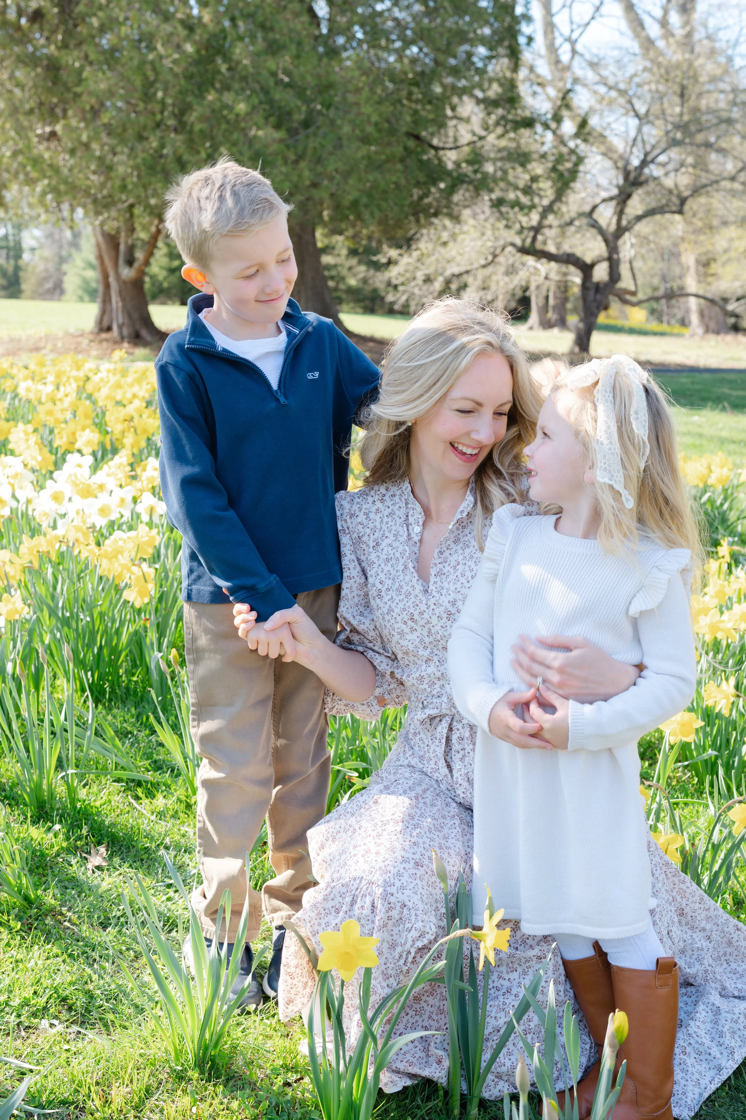 A woman with blonde hair, wearing a floral dress, is kneeling in a field of yellow daffodils and holding hands with a young girl in a cream-colored dress with puffed sleeves and brown boots. Another young boy with blonde hair, wearing a blue jacket a