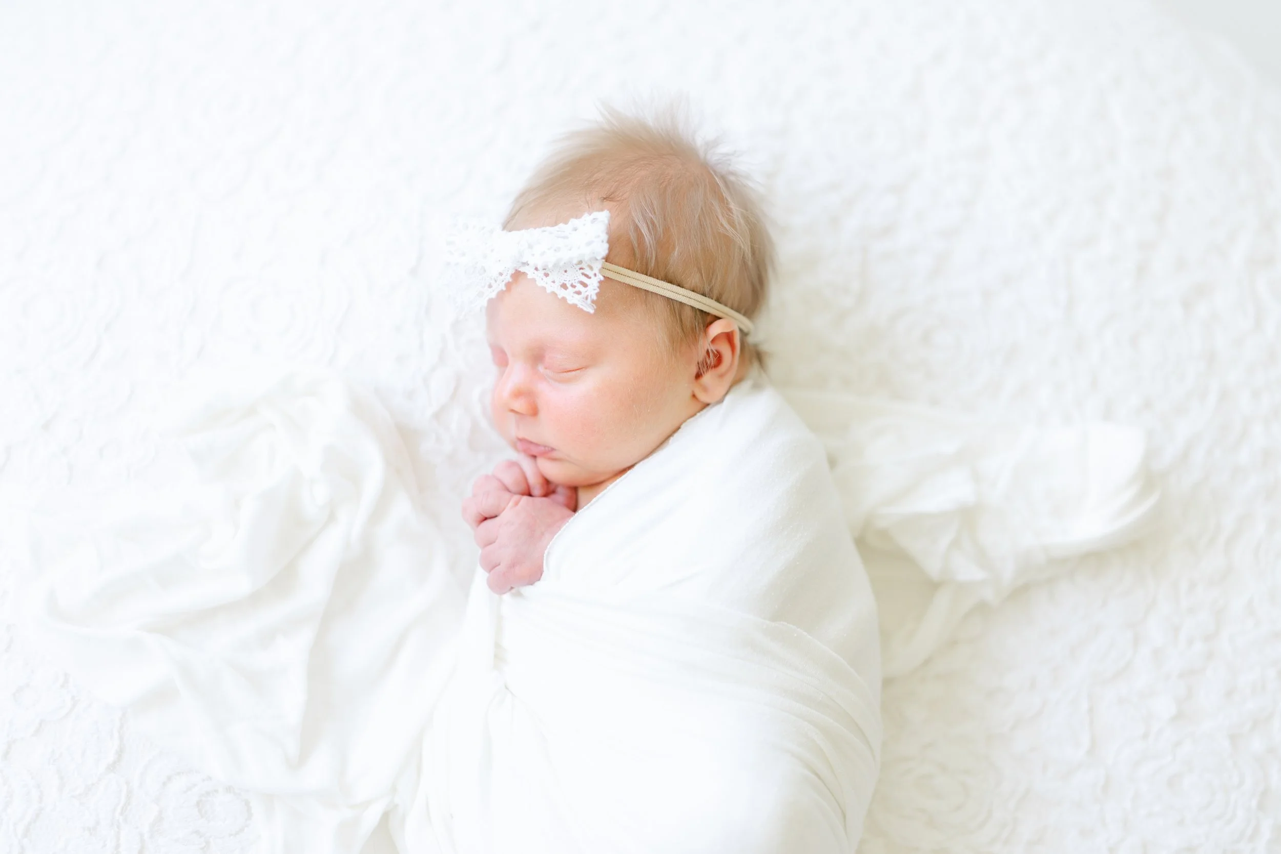 A peacefully sleeping baby with a headband and lace decoration on a soft, all-white bed or blanket.