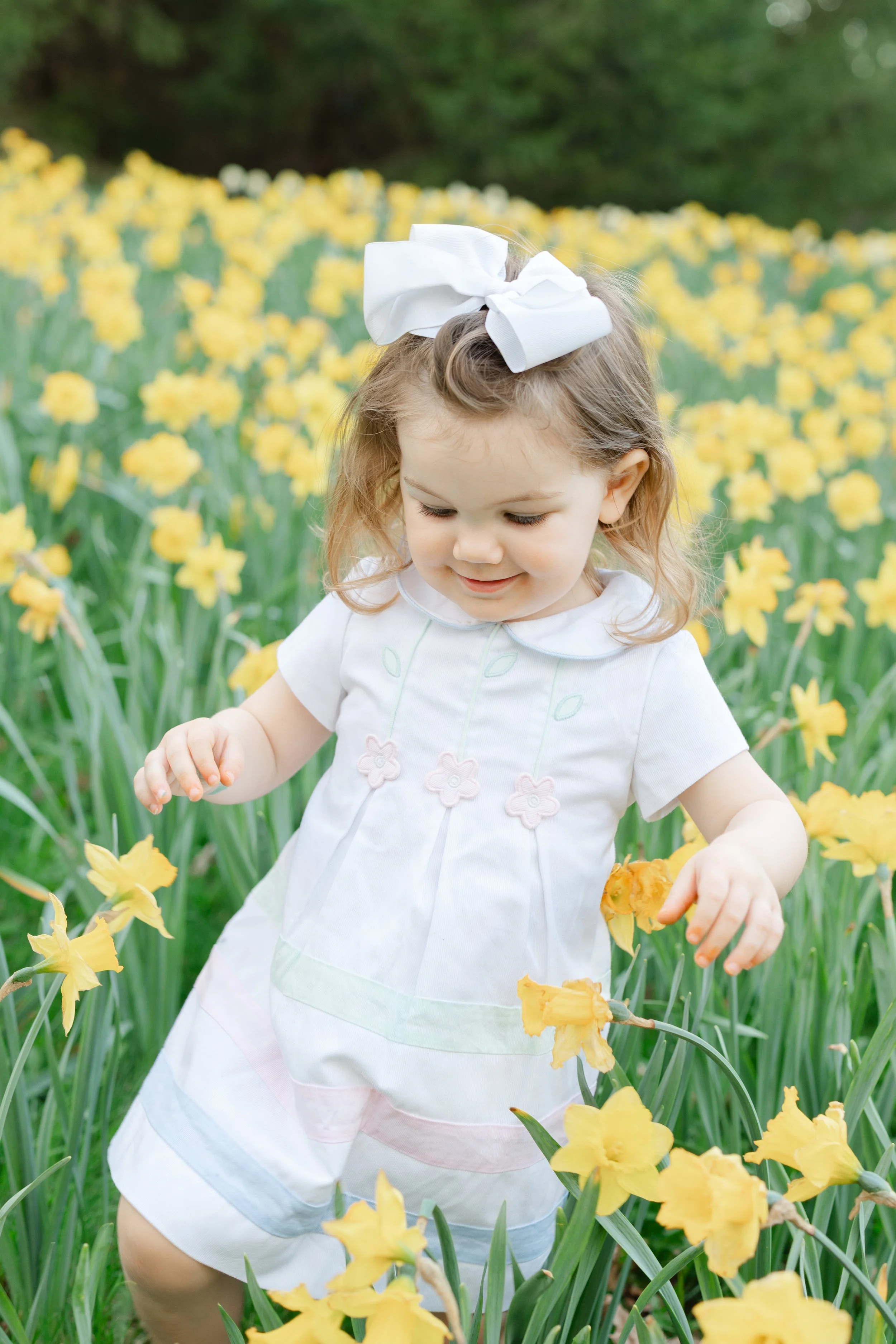 A young girl with light brown curly hair, wearing a white dress with pastel pink and green accents, and a large white bow in her hair, stands in a field of yellow flowers with a blurred green forest background.