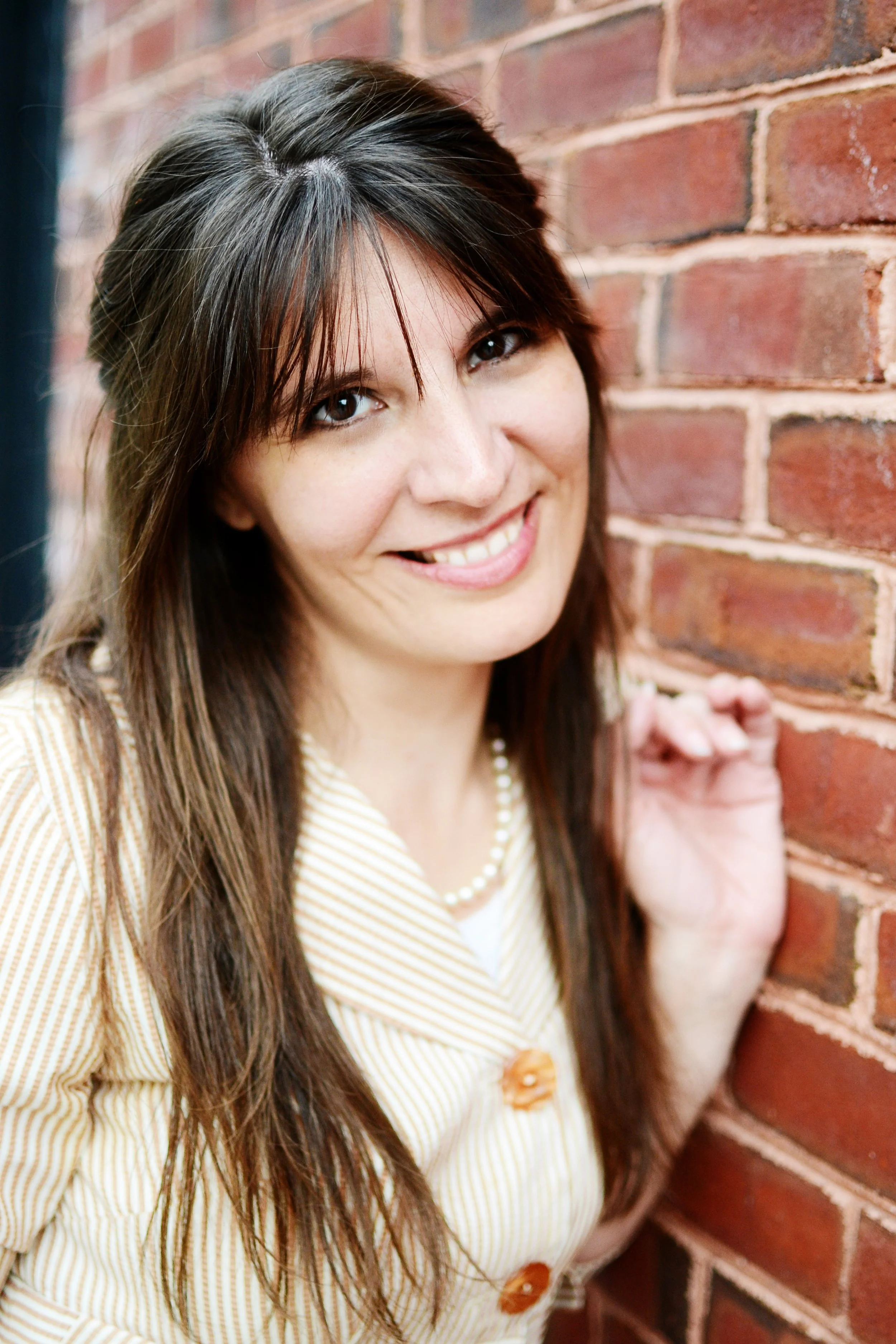 A woman with long brown hair and brown eyes smiling while leaning against a red brick wall.