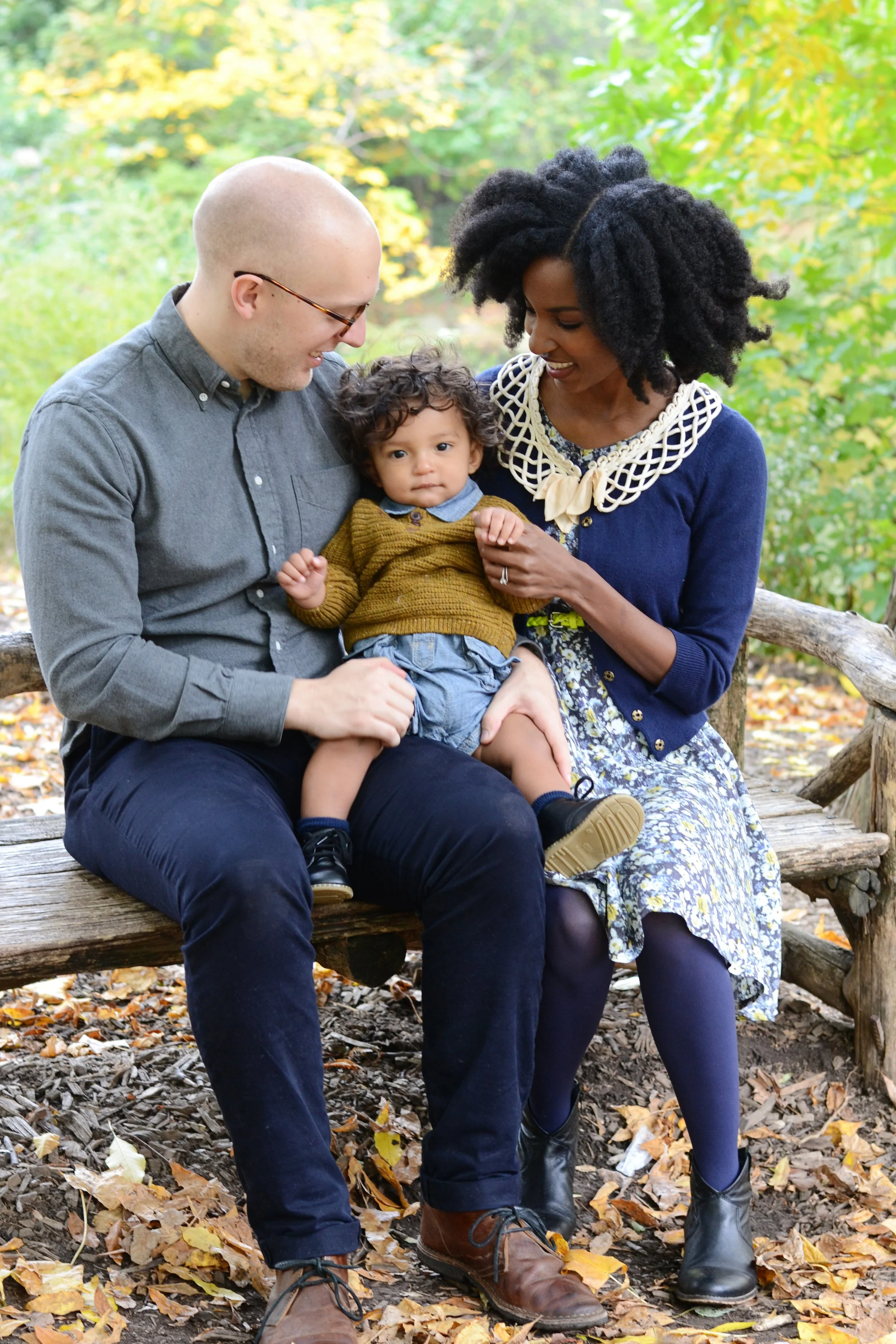 A family of three sitting on a wooden bench outdoors surrounded by fall leaves and trees. The man has a shaved head, glasses, a gray shirt, and dark pants, holding a young boy with curly hair in a mustard sweater and blue shorts. The woman has large,