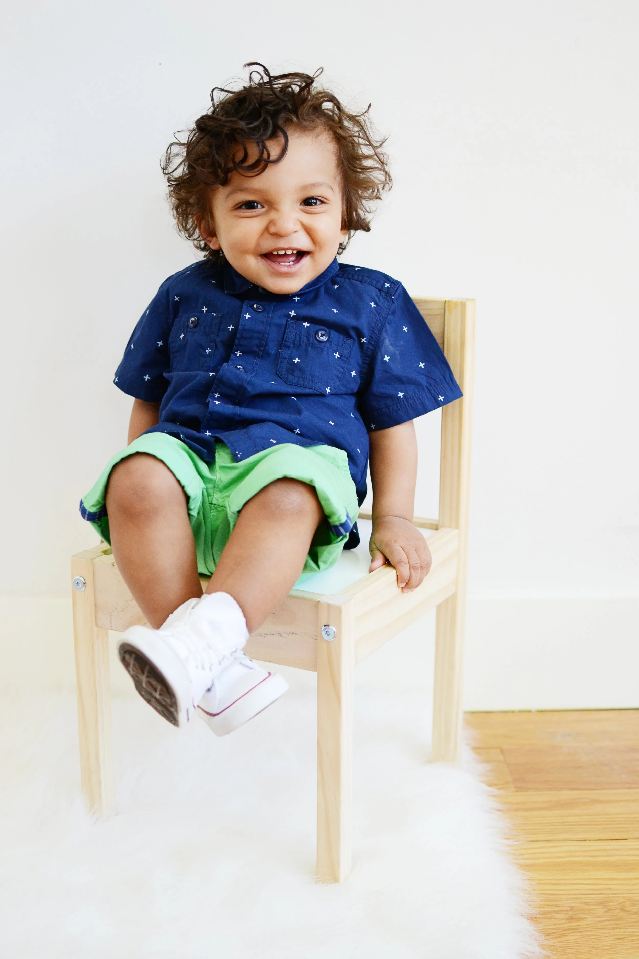 A young boy with curly hair smiling and sitting on a small wooden chair against a plain white wall.