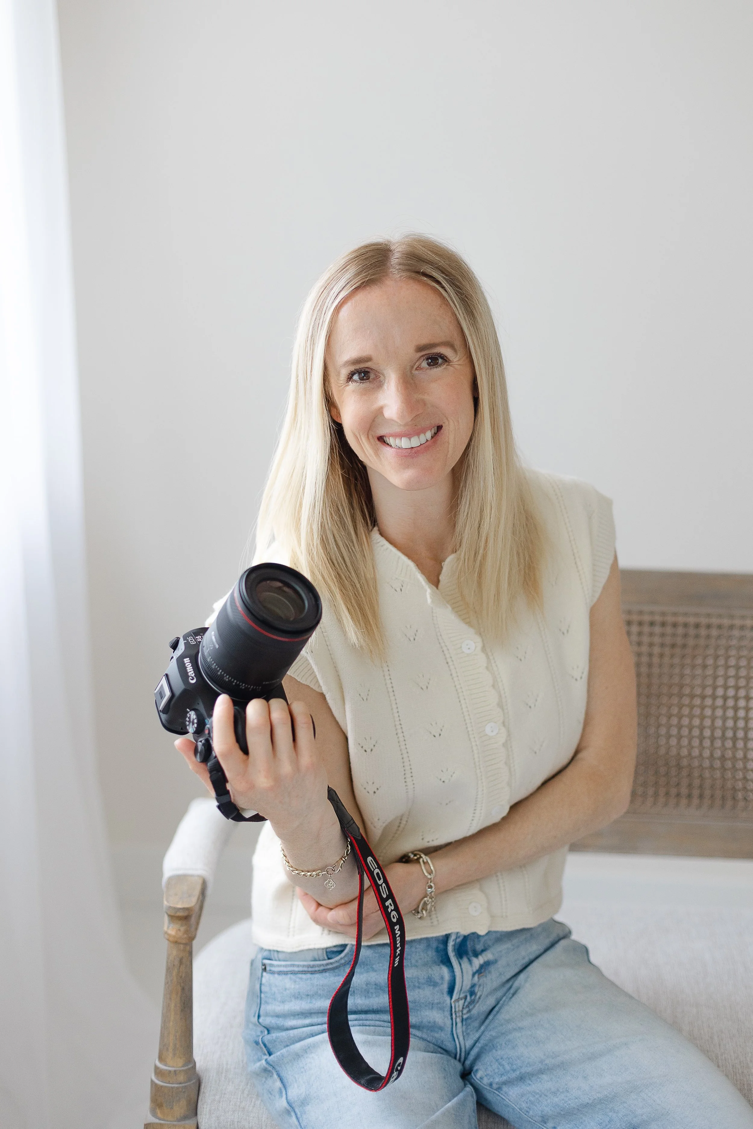 A smiling woman with blonde hair sitting on a wooden chair, holding a Canon camera lens in her hand, wearing a cream-colored sleeveless top and light blue jeans, in a minimalistic room with a plain white wall and a wicker chair in the background.