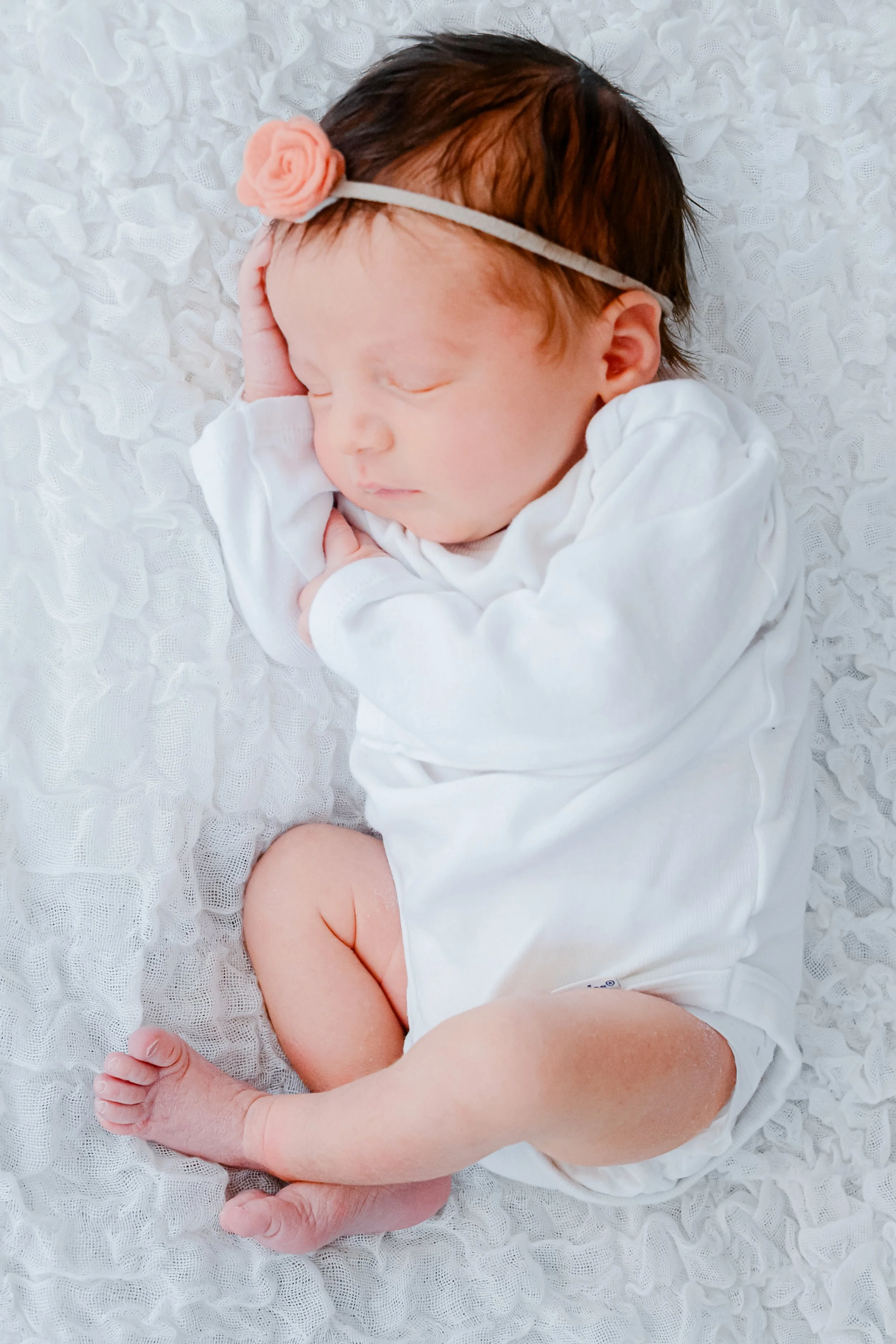 A sleeping newborn baby with a headband and a small pink flower, lying on a white textured blanket, dressed in a white onesie with a short sleeve.
