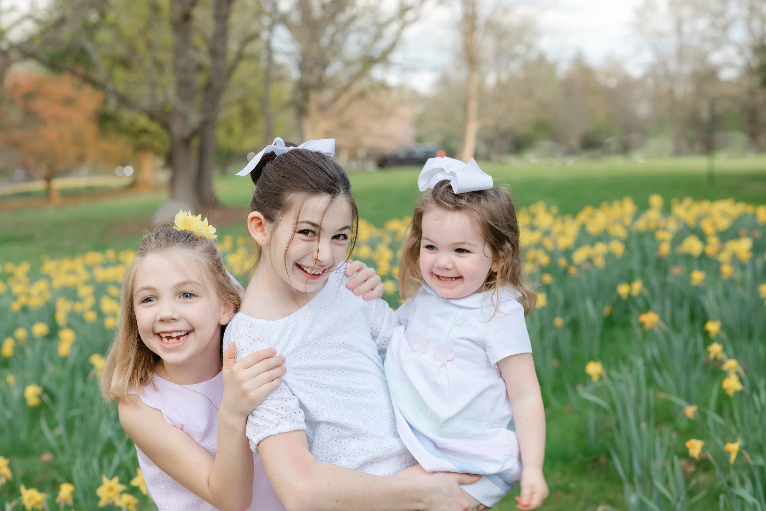 Four young girls happily hugging outdoors in a park filled with yellow flowers, trees, and green grass on a bright day.