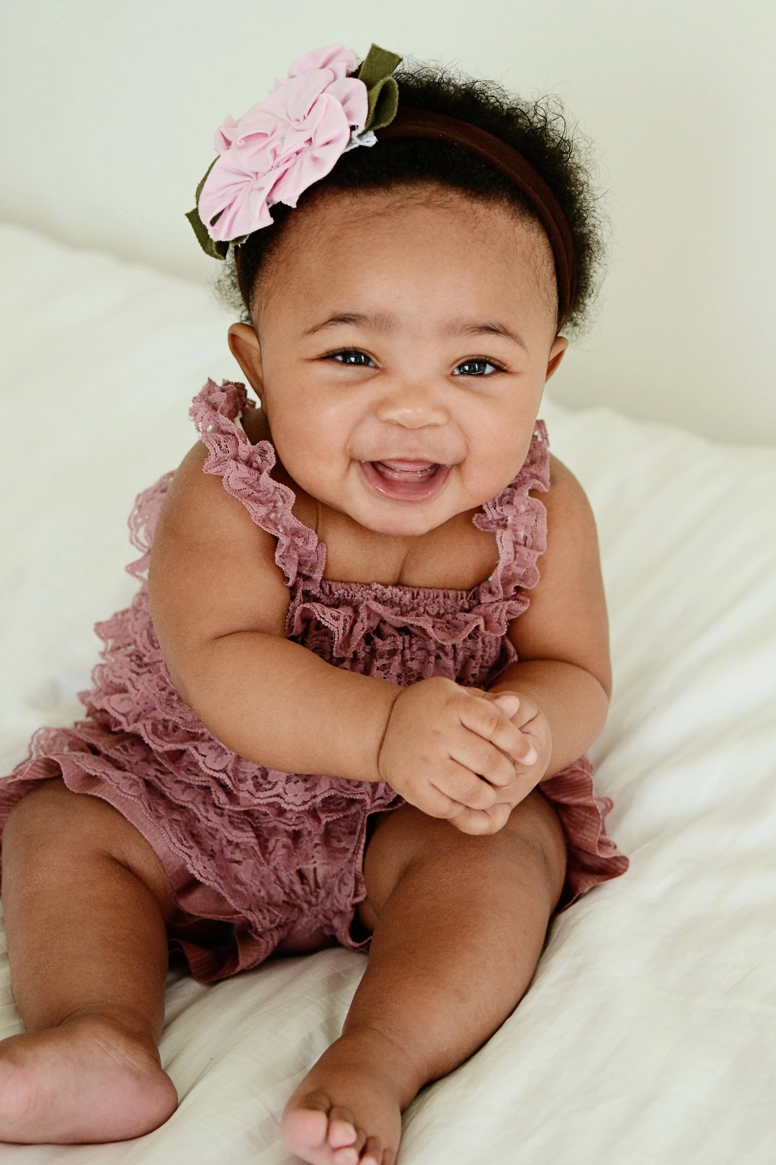 A smiling baby girl with curly hair, wearing a pink ruffled outfit and a pink flower headband on a cream-colored bed.
